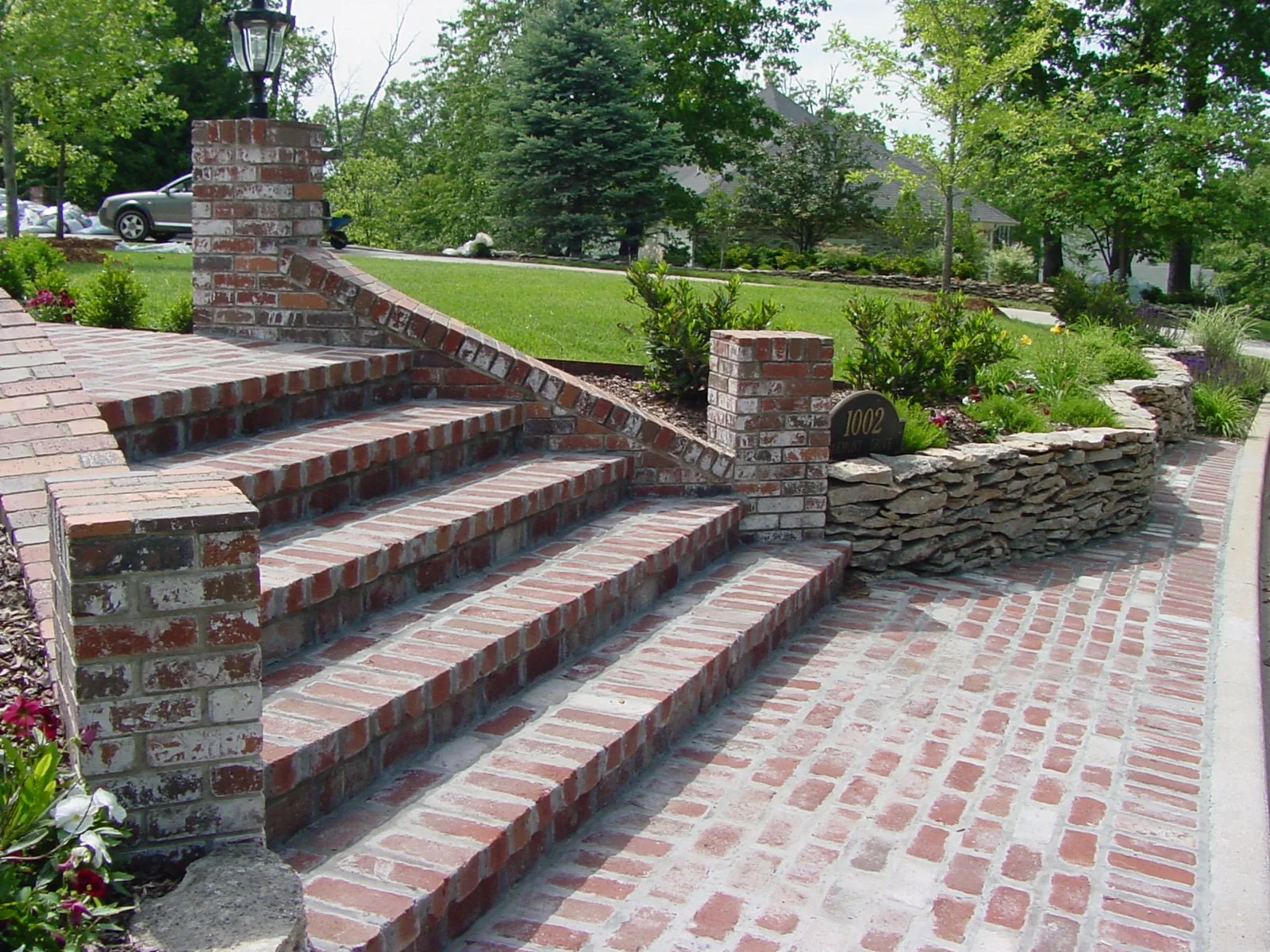 Brick staircase leading to a garden with a brick and stone retaining wall, green trees, and a suburban house in the background.