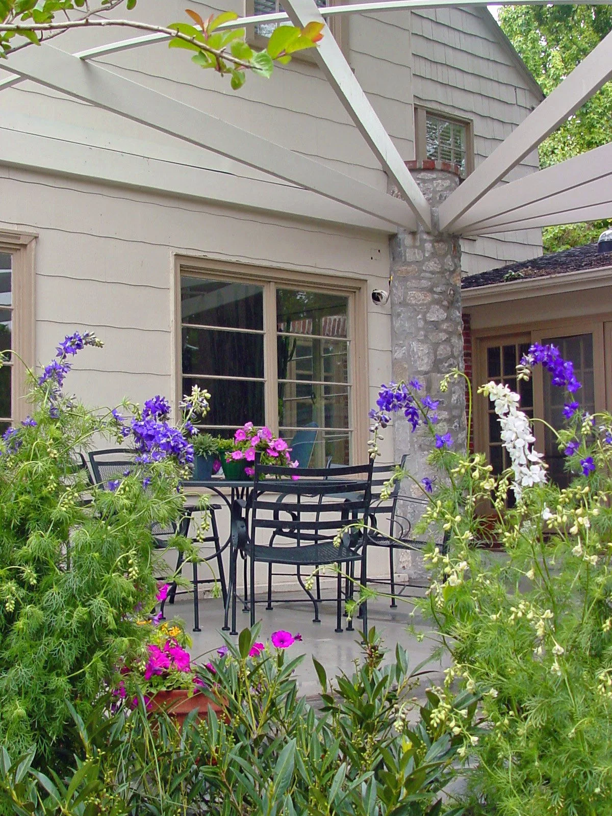 A backyard patio with black metal furniture including a round table and chairs, decorated with pink and purple flowers in pots. The background shows a house with beige siding, a stone chimney, and a window, with lush greenery and blooming plants in t