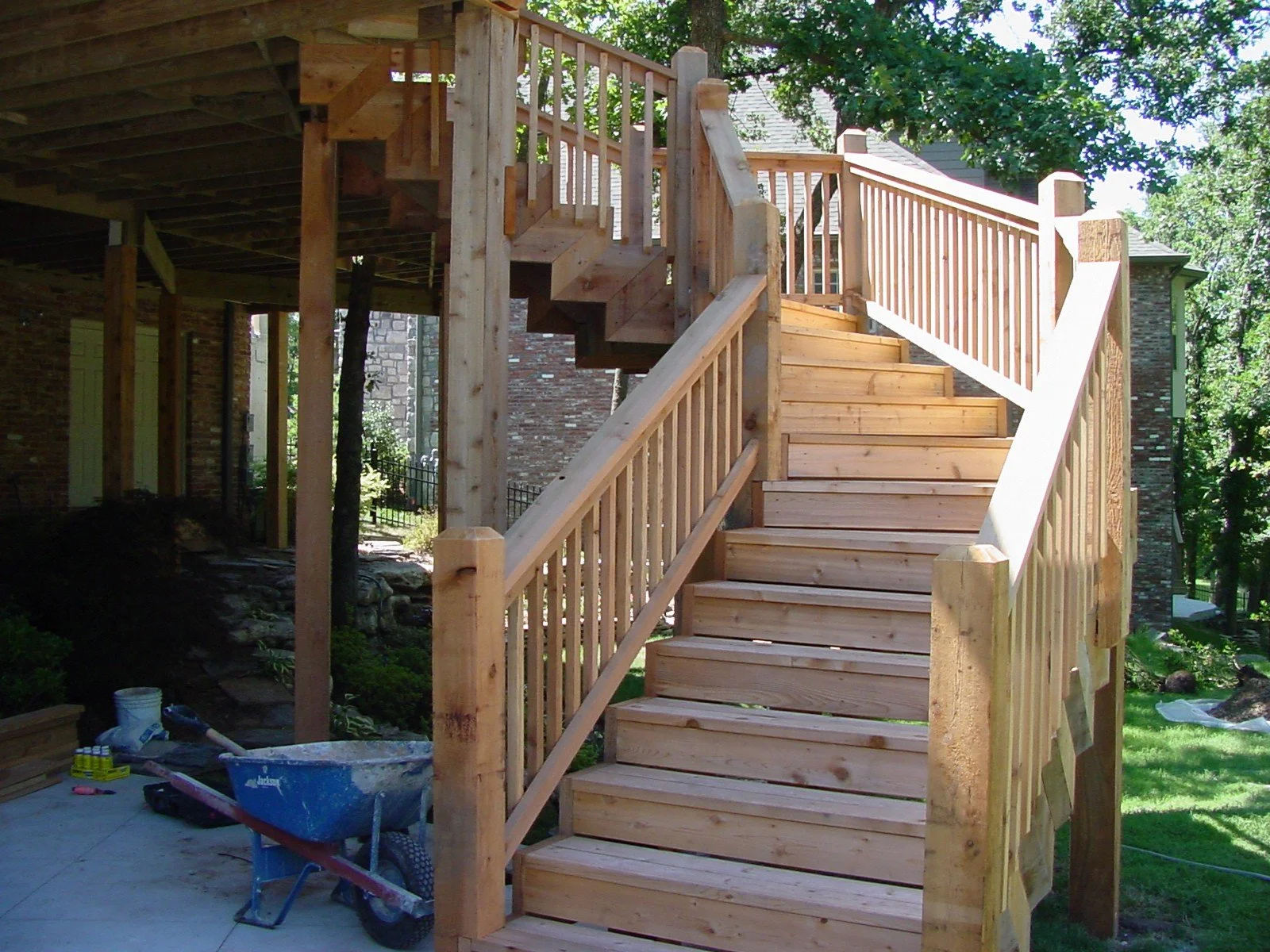 Wooden outdoor staircase under construction with new handrails and stairs, surrounded by green trees and neighboring houses.