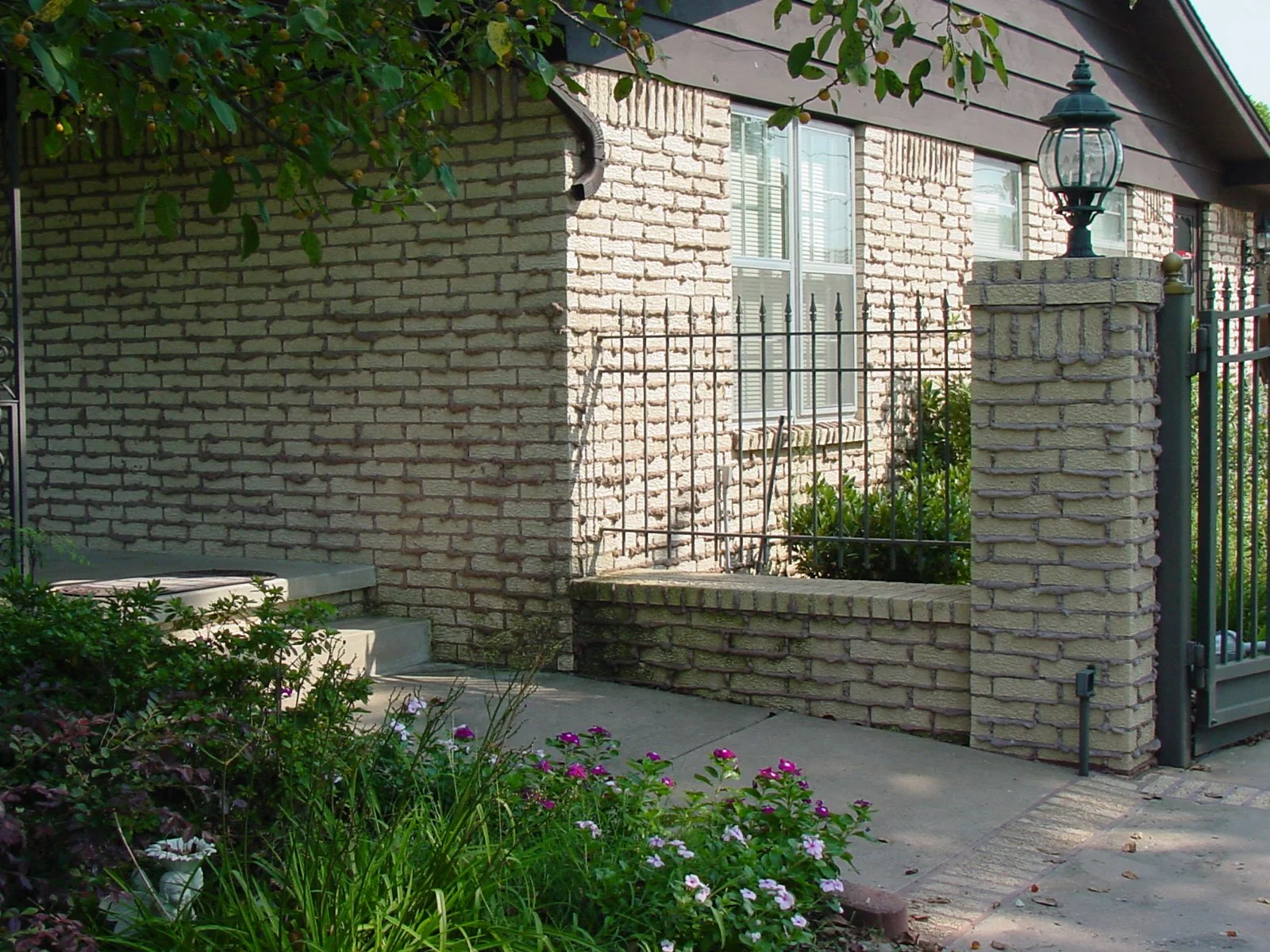 A brick house exterior with a small front yard, featuring a concrete step, green plants, and pink flowers. A lamp is mounted on a brick pillar next to a black metal gate.