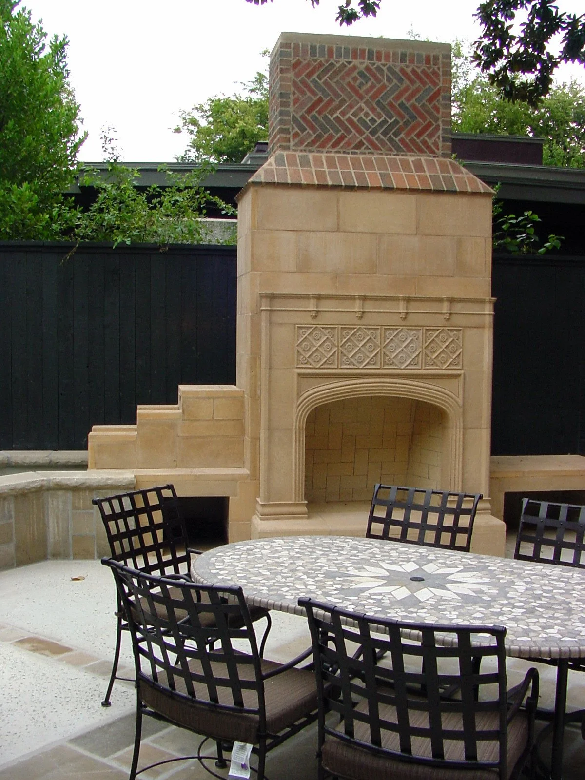 Outdoor patio area with a stone fireplace, a round mosaic table, and black metal chairs. Behind the fireplace, there's a black fence and some green trees.