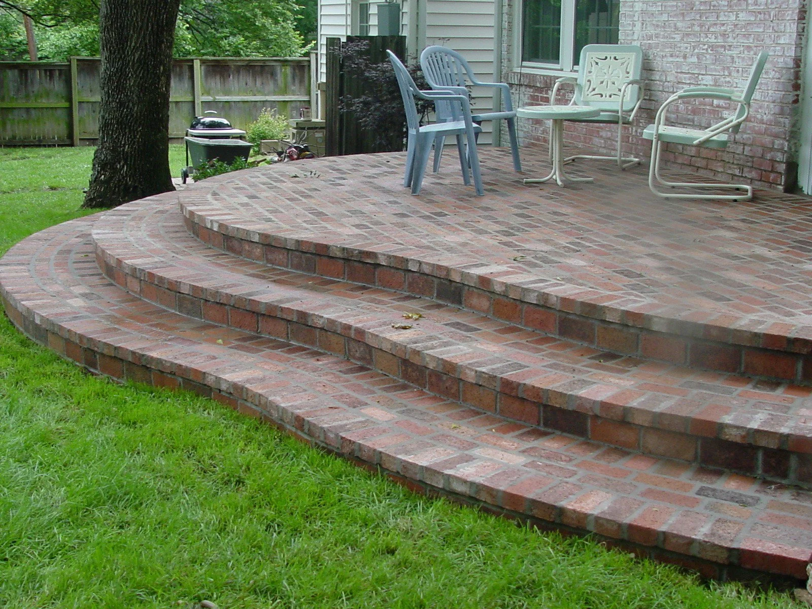 Brick patio with curved steps leading up to a house, outdoor furniture including chairs and a small table, surrounded by a grassy yard and a wooden fence.