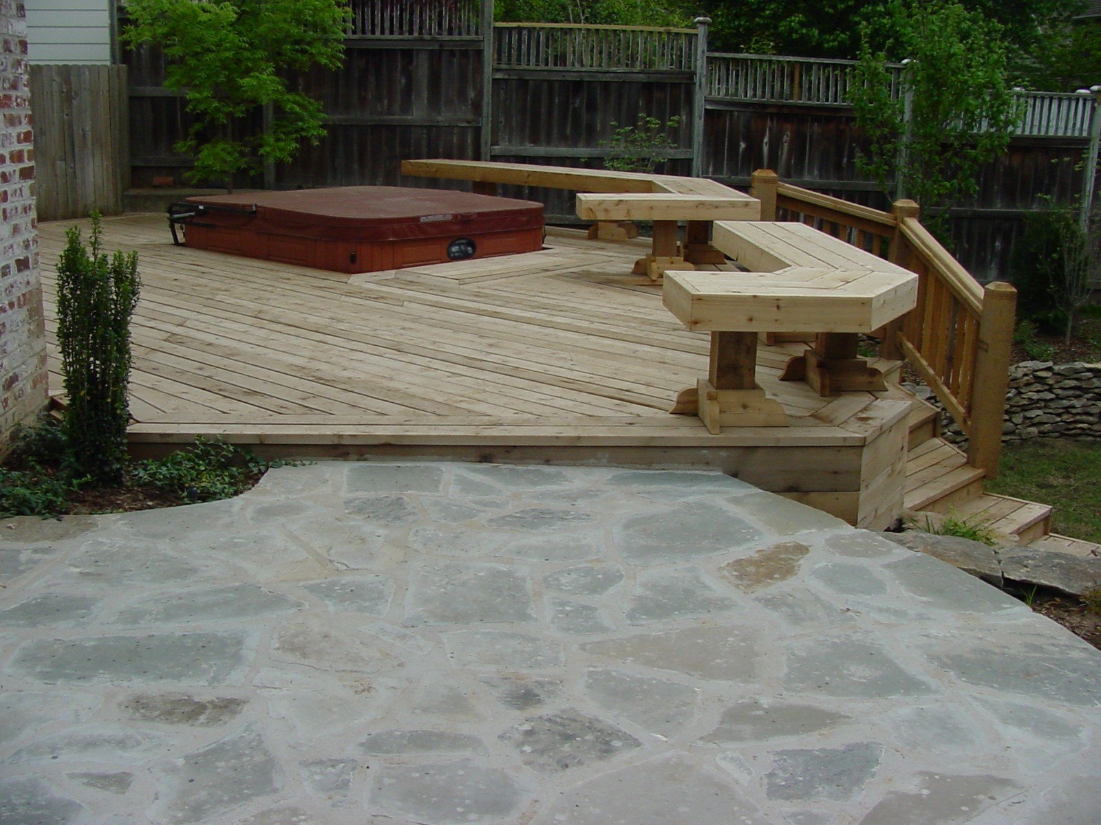 A backyard wooden deck with railing, stairs, and a hot tub, bordered by plants and trees, with a stone patio in the foreground.