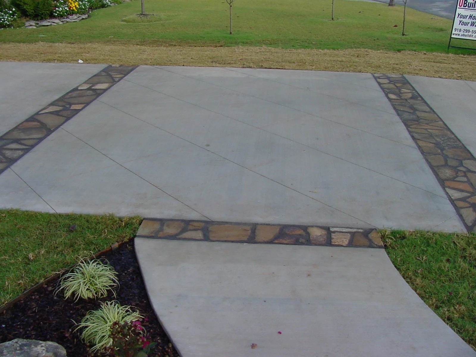 Concrete sidewalk with decorative stone borders, grass and plants along edges, small trees in background, and a sign on the right side.