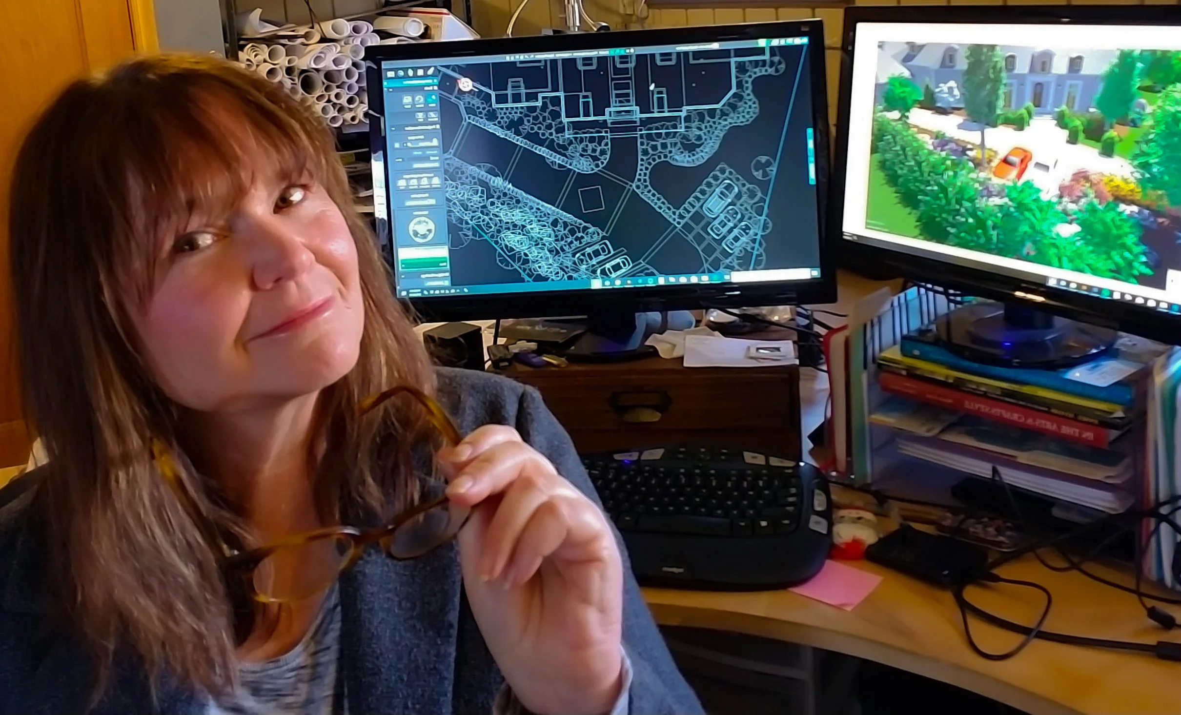 A woman with long, reddish-brown hair and glasses holding her glasses in her right hand, sitting at a cluttered desk with two computer monitors. One monitor displays a detailed digital map or blueprint, and the other shows a colorful 3D landscape or garden scene. There are papers, books, and other items on the desk and shelves in the background.