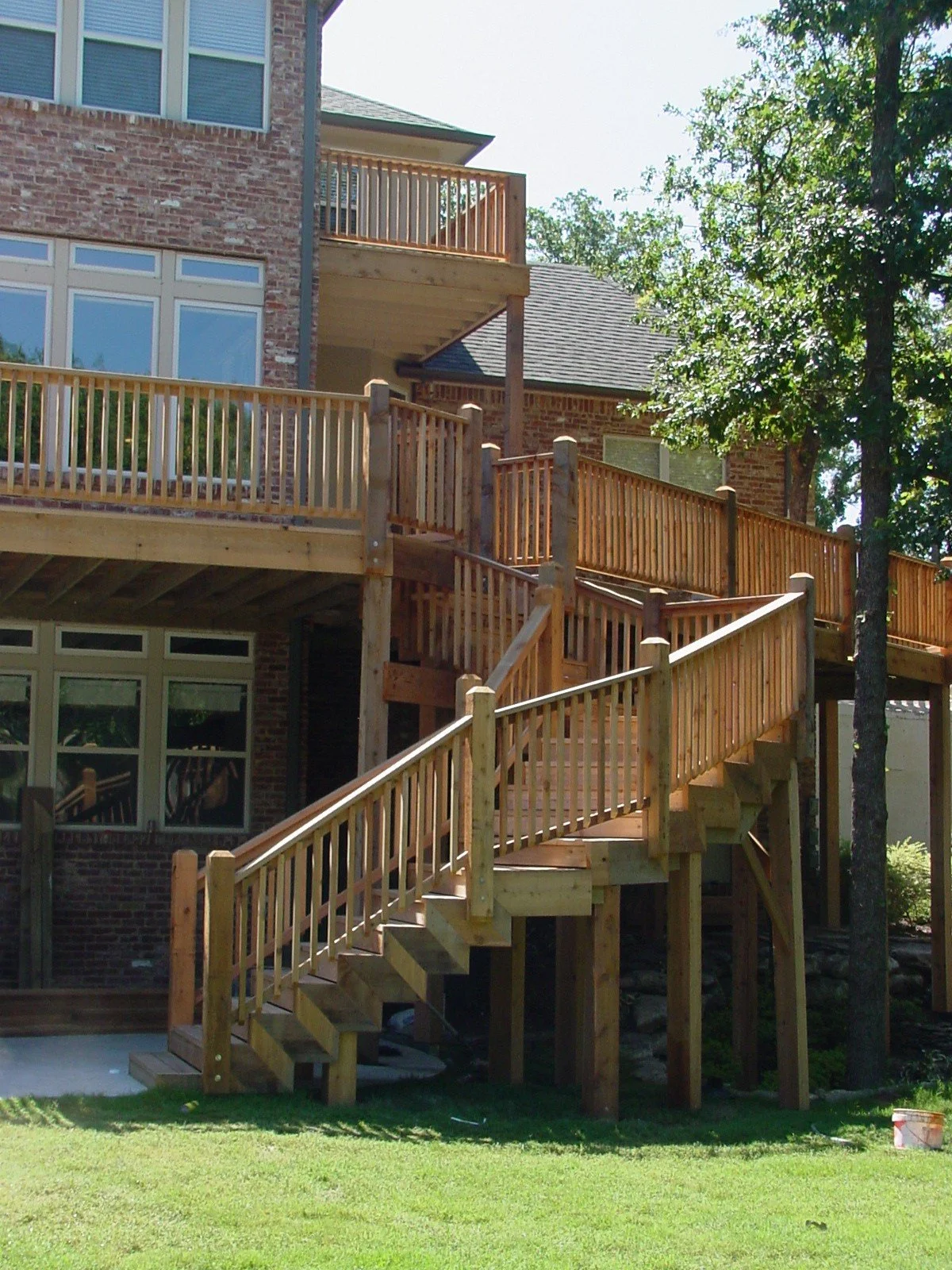 Wooden stairs and balcony railings attached to the back of a brick house, with trees and a grassy yard in the background.