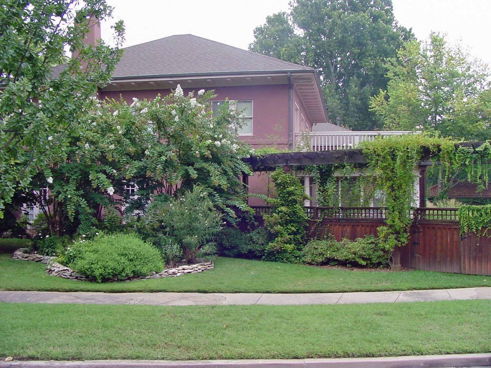 A two-story house with a purple exterior, surrounded by a lush green garden with various trees and shrubs, and a wooden fence with climbing plants.
