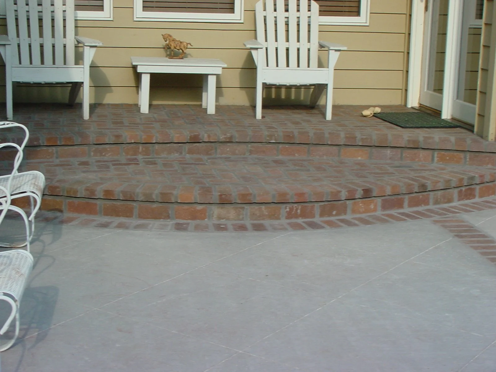 A brick patio with two white wooden chairs and a small white table between them, a pair of shoes near the door, and a brick step leading up to a yellow house with a door and window