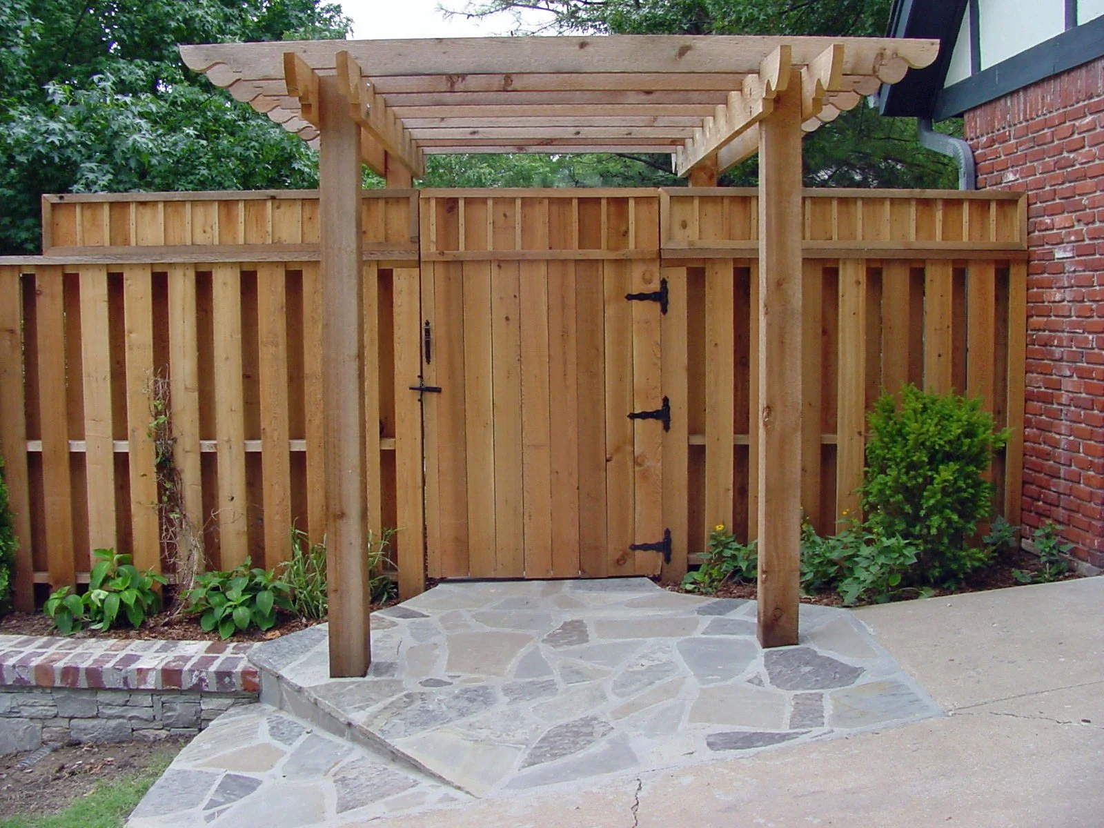 A wooden fence with a gate and a pergola above it, adjacent to a brick house wall. There are plants and small bushes on either side of the gate, and a stone pathway leading to the gate.