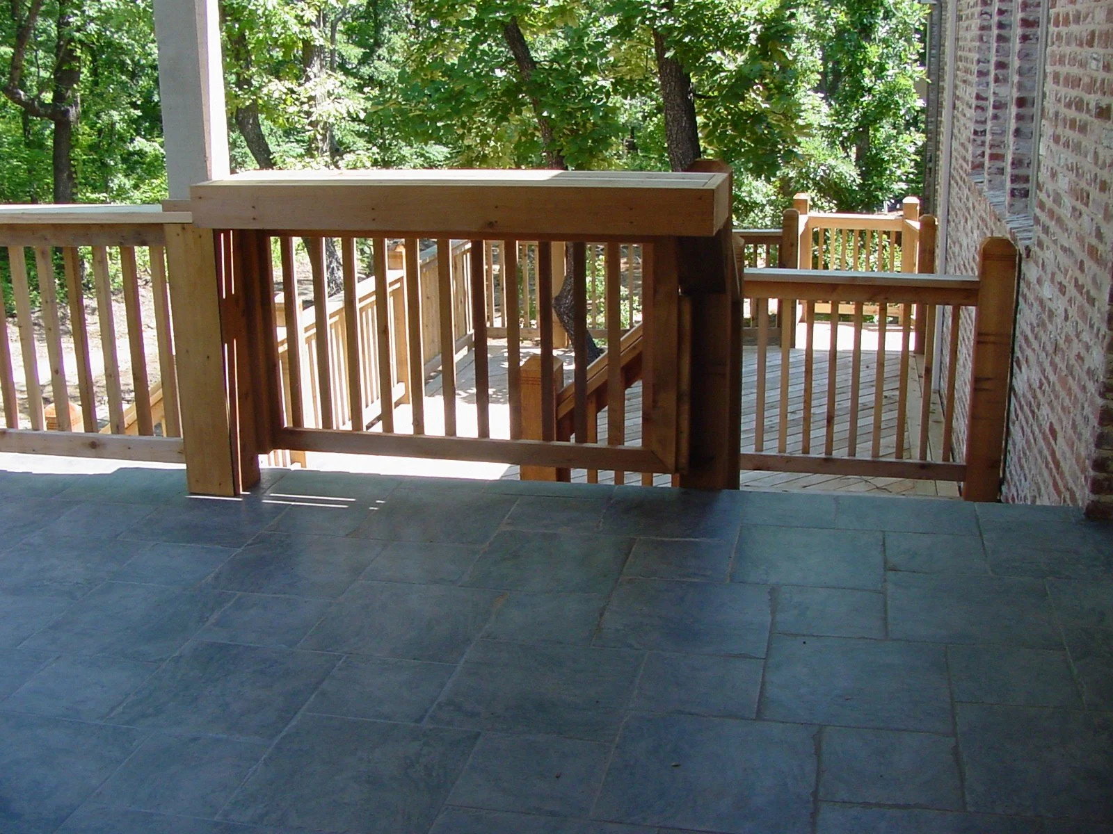 A wooden balcony with a railing and stairs leading down to a wooded area outside, with trees and greenery in the background.