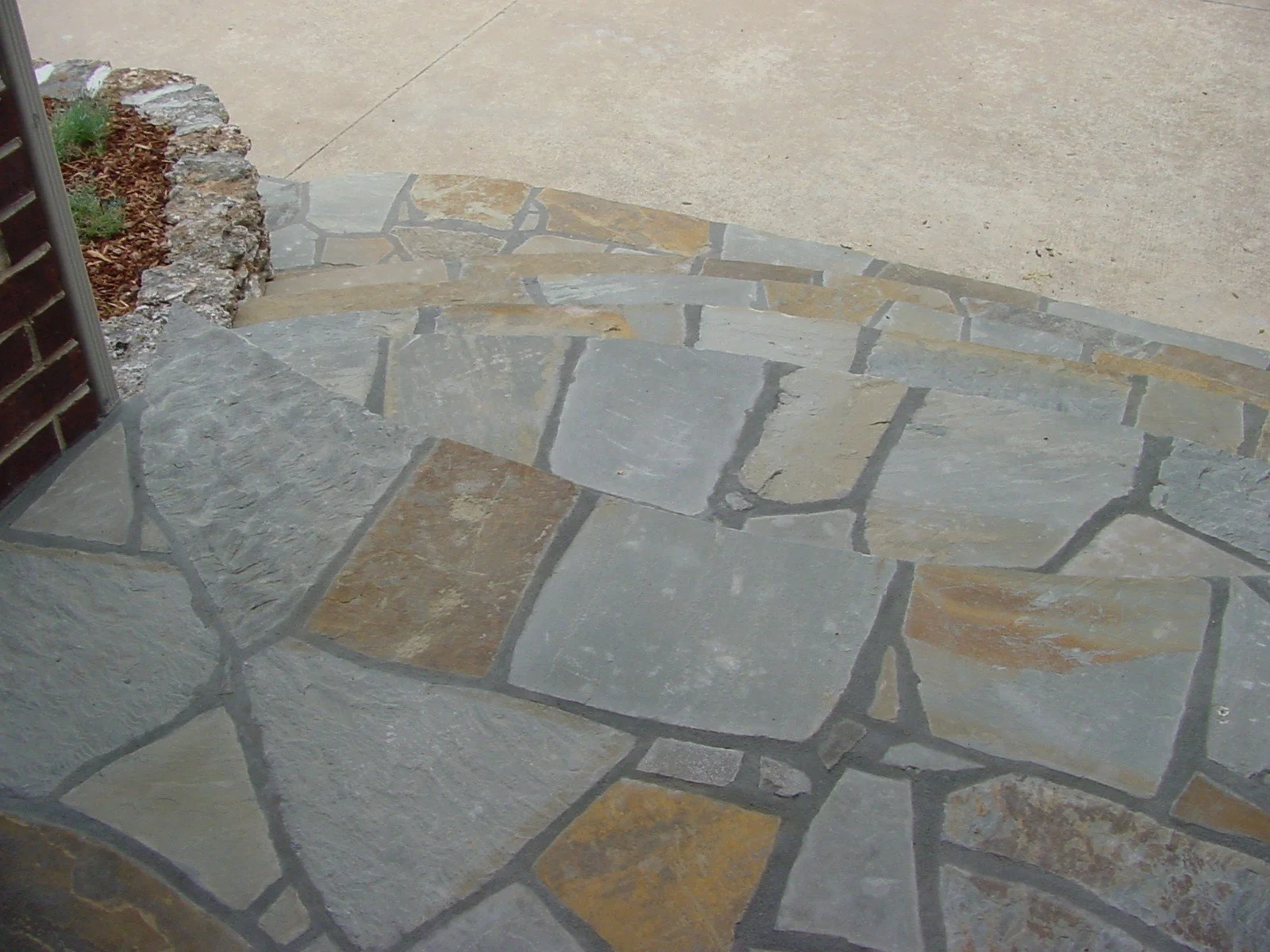 Close-up of a stone and concrete porch with a small garden bed on the left side, featuring a brick wall and some green plants.
