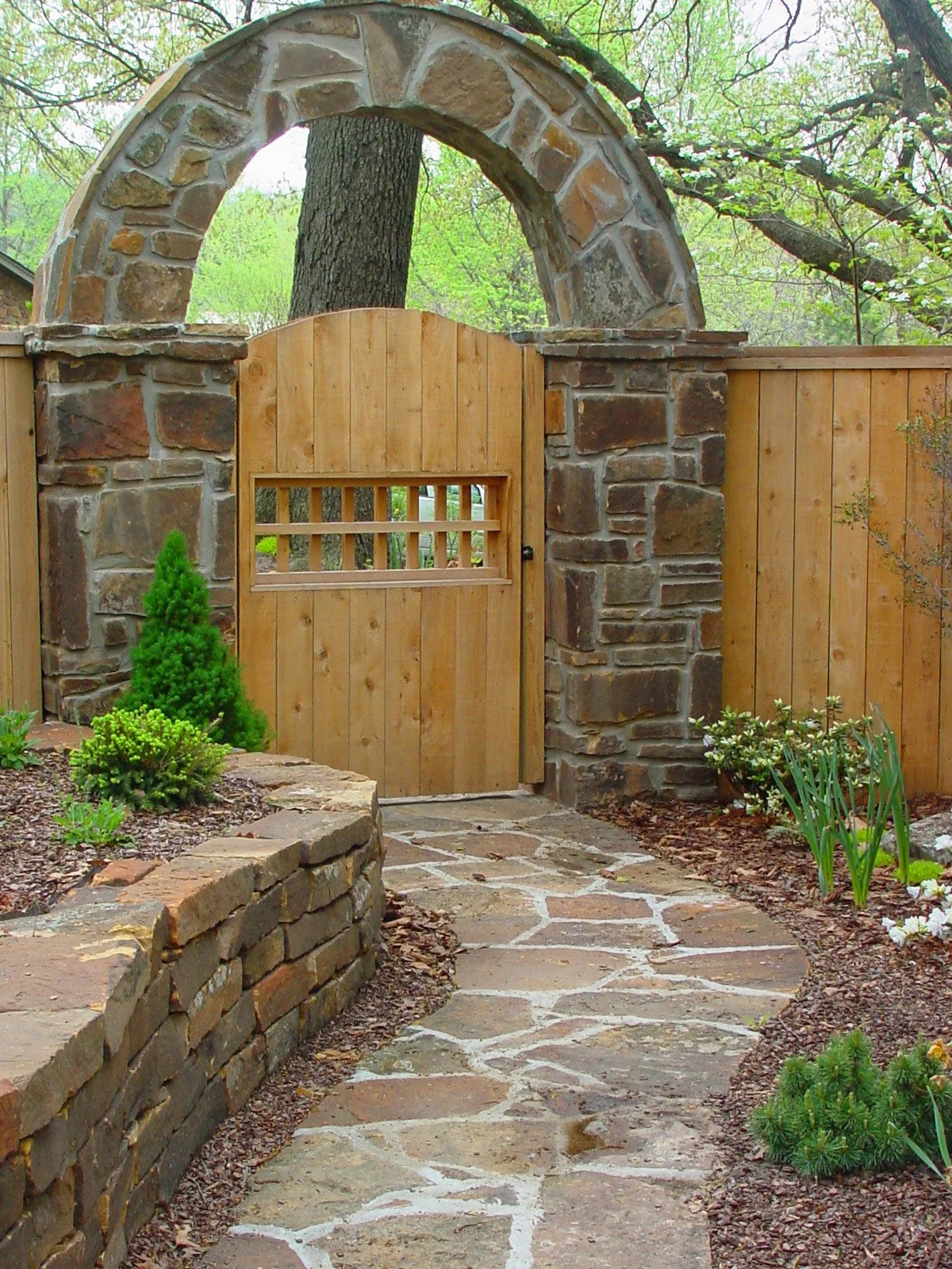 A garden gate with a stone archway and a wooden door, surrounded by a stone border, plants, and a stone pathway.