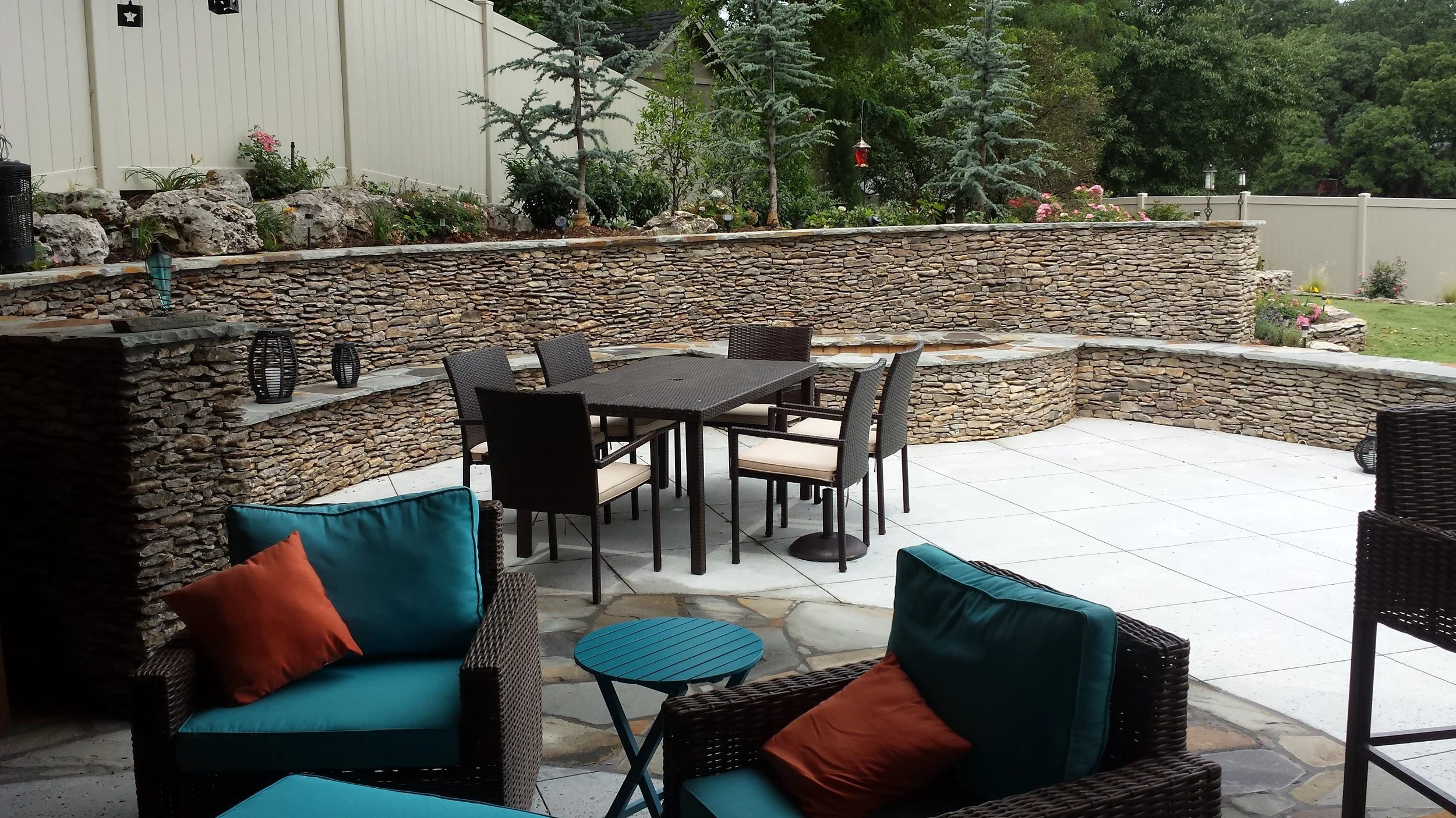 A backyard patio with outdoor furniture, a stone retaining wall, potted decorations, and a landscaped garden with trees and plants.