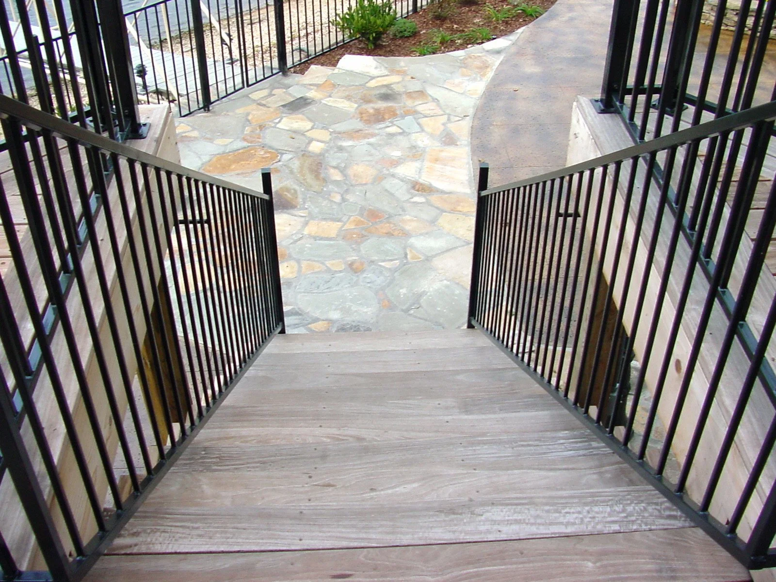 View looking down a small balcony with black metal railing, wooden floor, and a stone patio below with a curved walkway and some green plants.