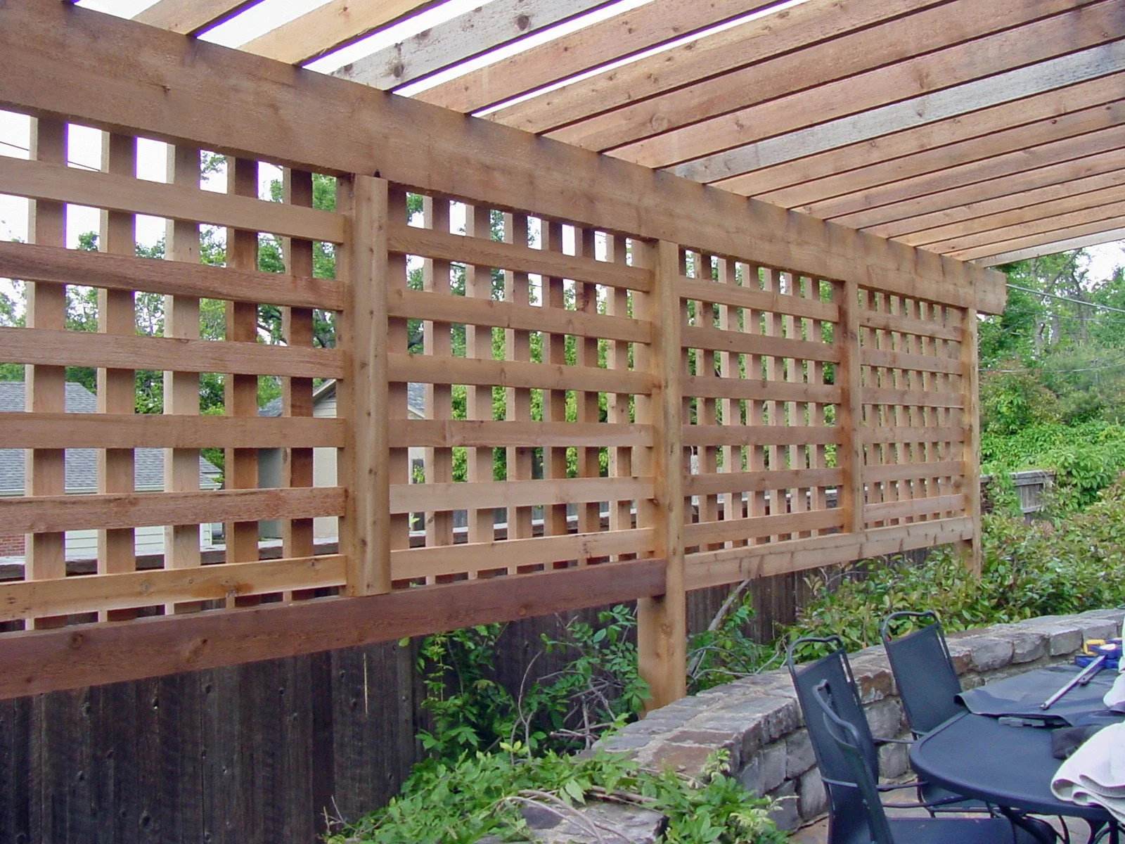 View of a partially constructed wooden privacy fence in a backyard, with a stone patio and outdoor chairs in the foreground, and trees and neighboring houses in the background.