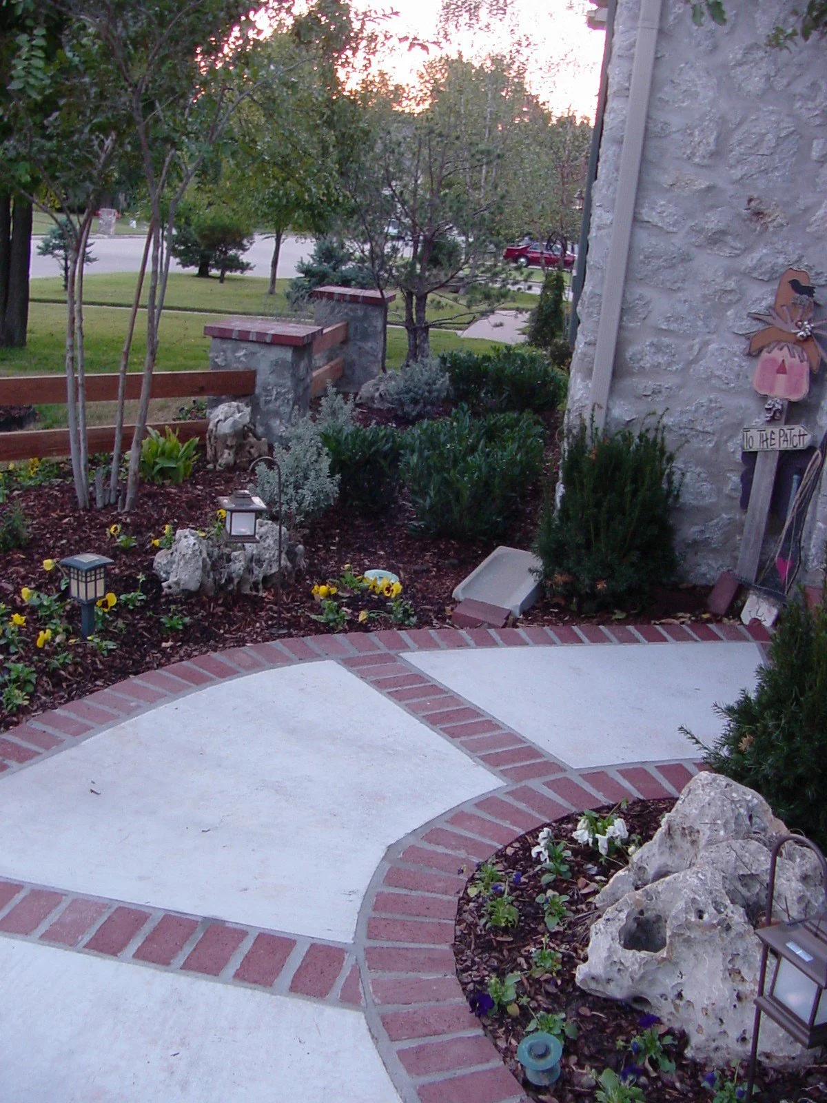 View of a landscaped front yard with a concrete and brick sidewalk, garden beds with plants and flowers, small decorative rocks, and a stone wall on the right side. There are some outdoor decorative items and lighting fixtures among the greenery, wit