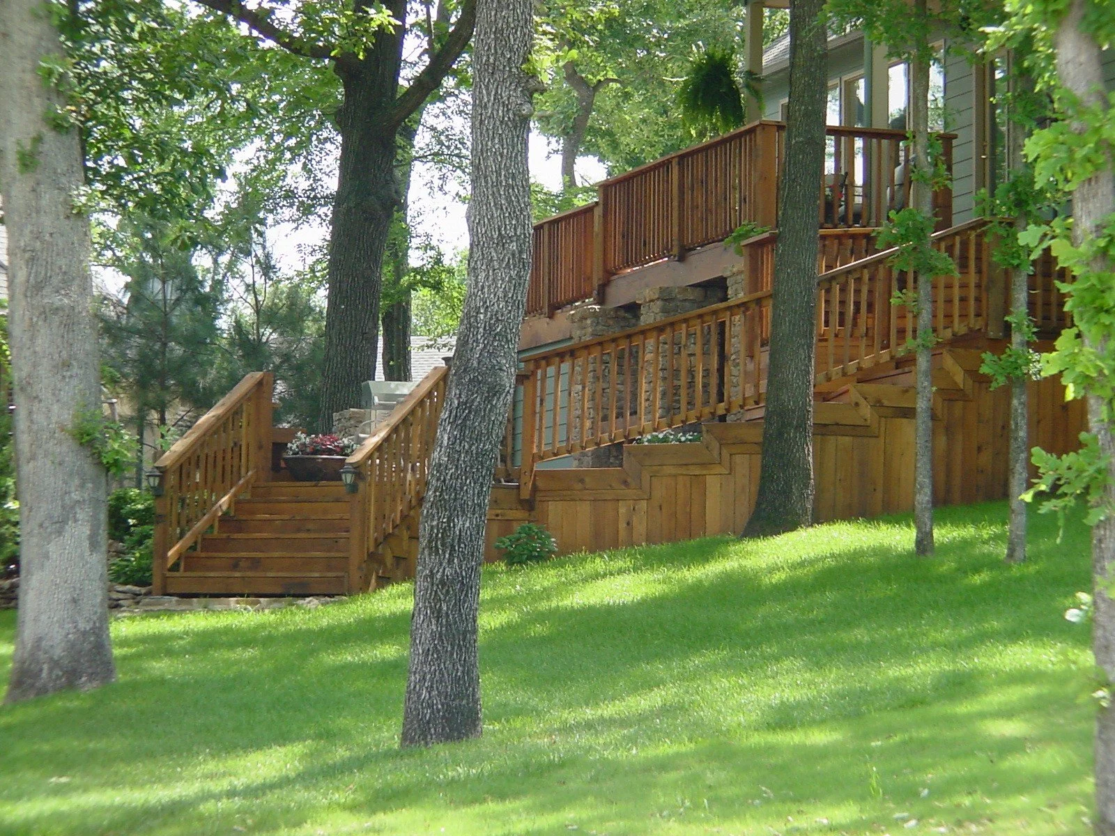 A wooden outdoor deck with stairs leading up to a house, surrounded by large trees and a well-maintained grassy yard.