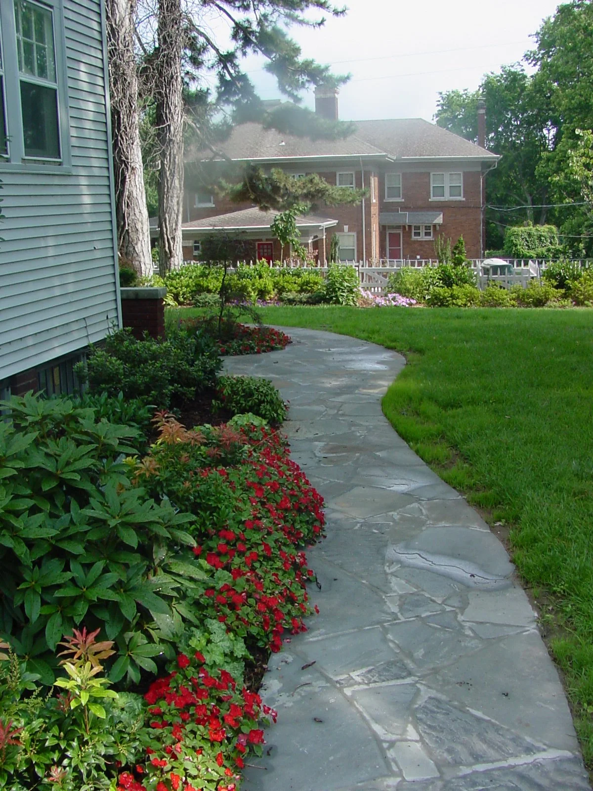 A stone pathway curves through a lush green garden with colorful flowers, leading to a fence and a brick house in the background, with trees and a neighboring building visible.