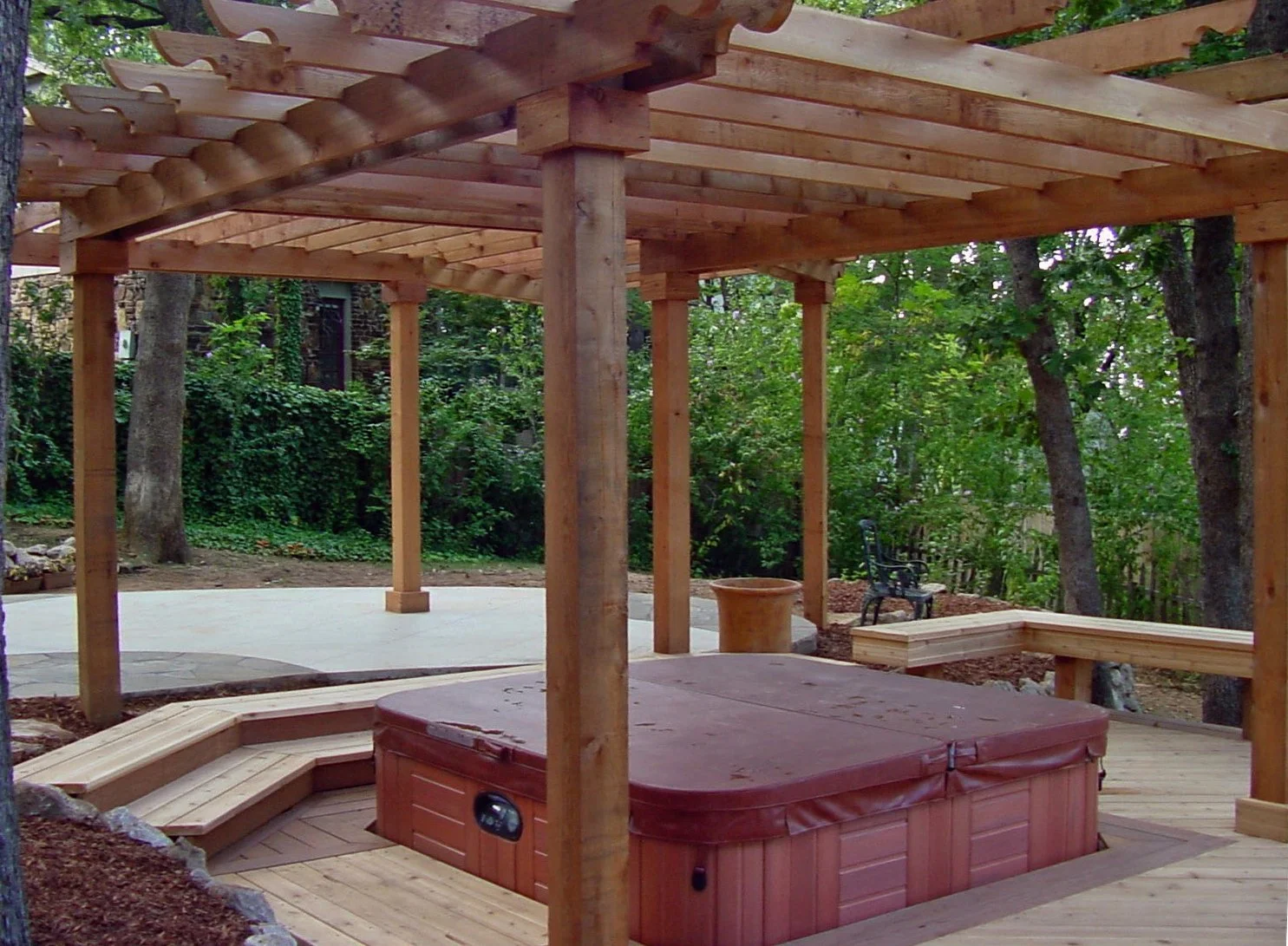 A partially built wooden deck with a hot tub, including a covered area with wooden support beams and a bench, in a backyard surrounded by trees and greenery.