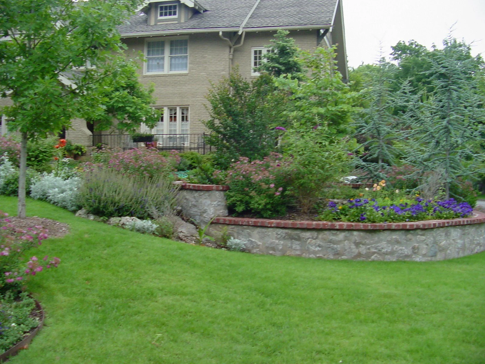 A landscaped front yard with a lush green lawn, various flowering plants, bushes, and trees, bordered by a stone and brick retaining wall in front of a beige house.