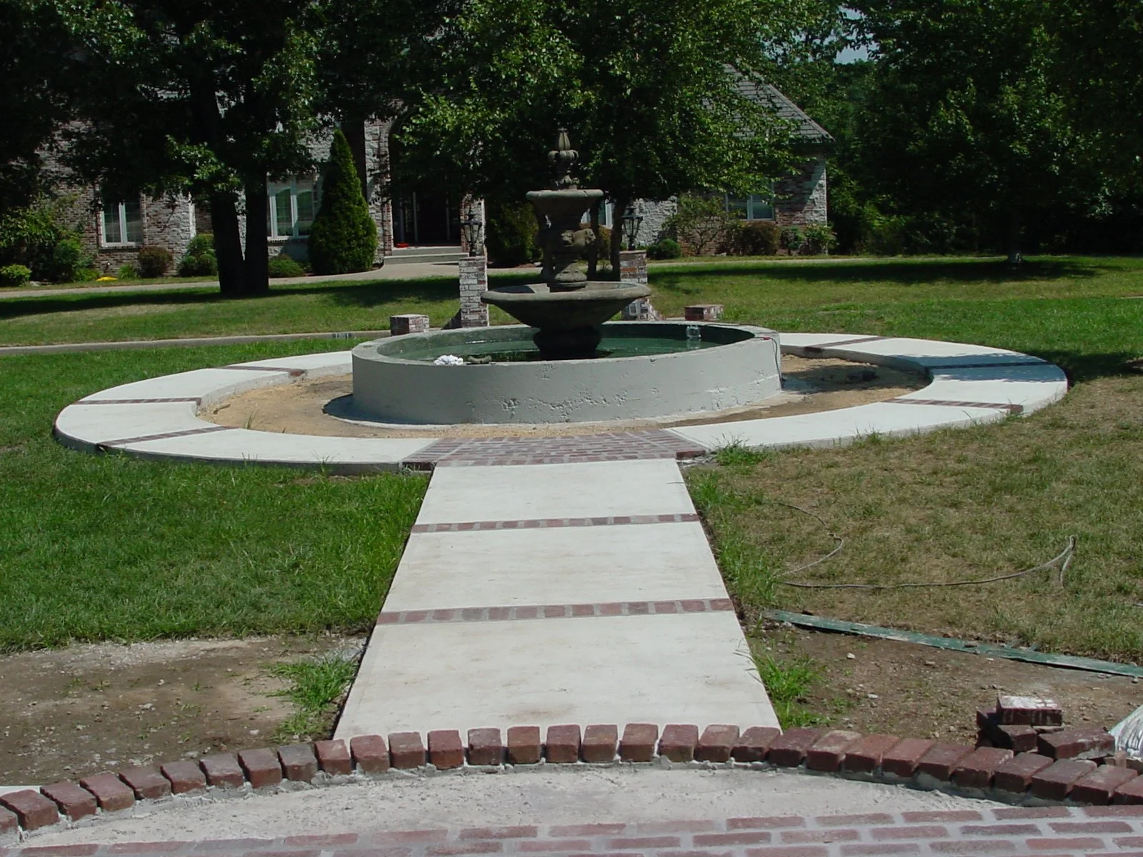 A landscaped front yard with a concrete pathway leading to a fountain in the center. The fountain has multiple tiers and is surrounded by a circular concrete border. In the background, there are trees and a house with brick walls and multiple windows