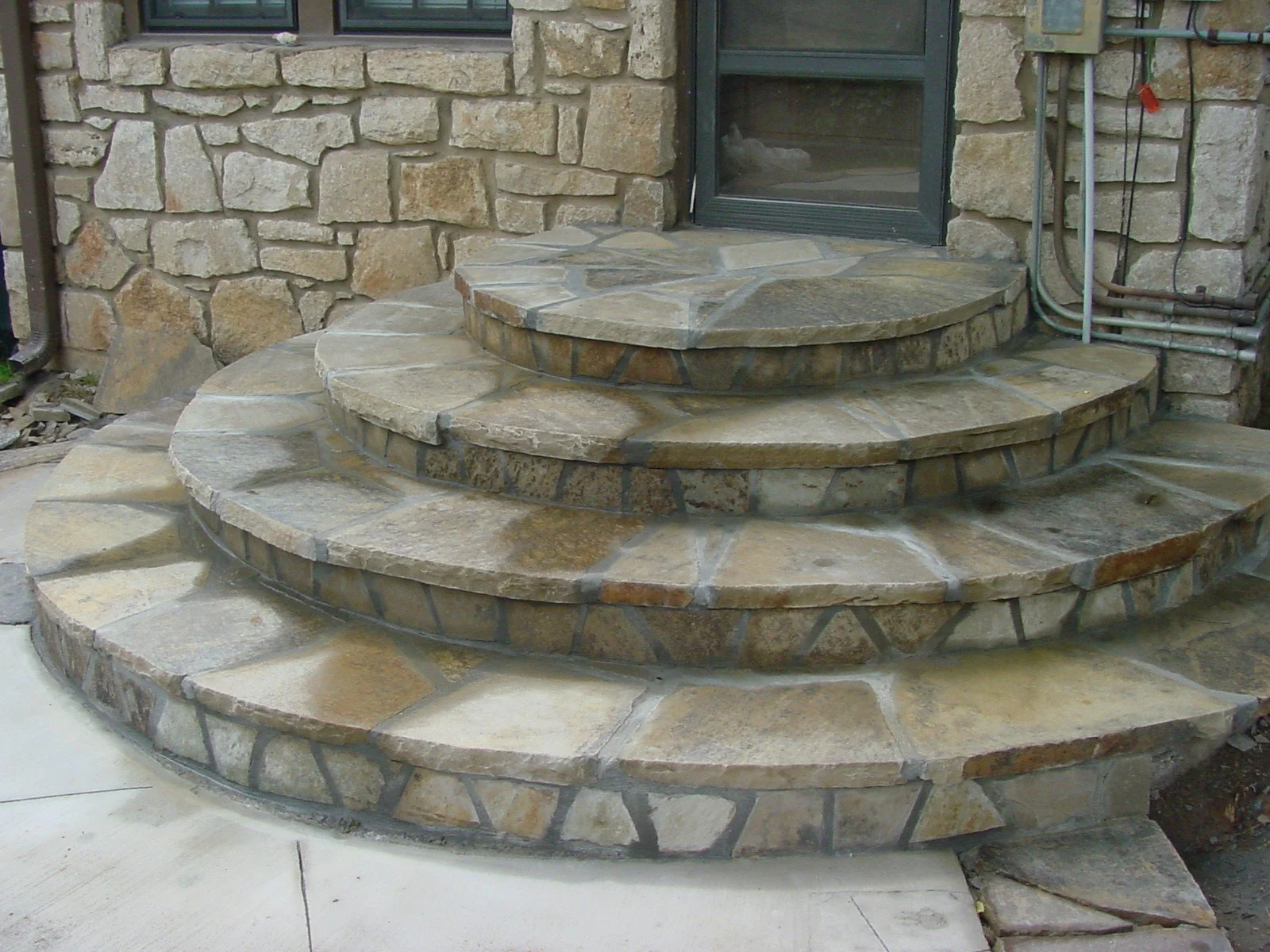 Stone steps leading to a glass door on a stone house exterior, with visible electrical wiring on the right side.