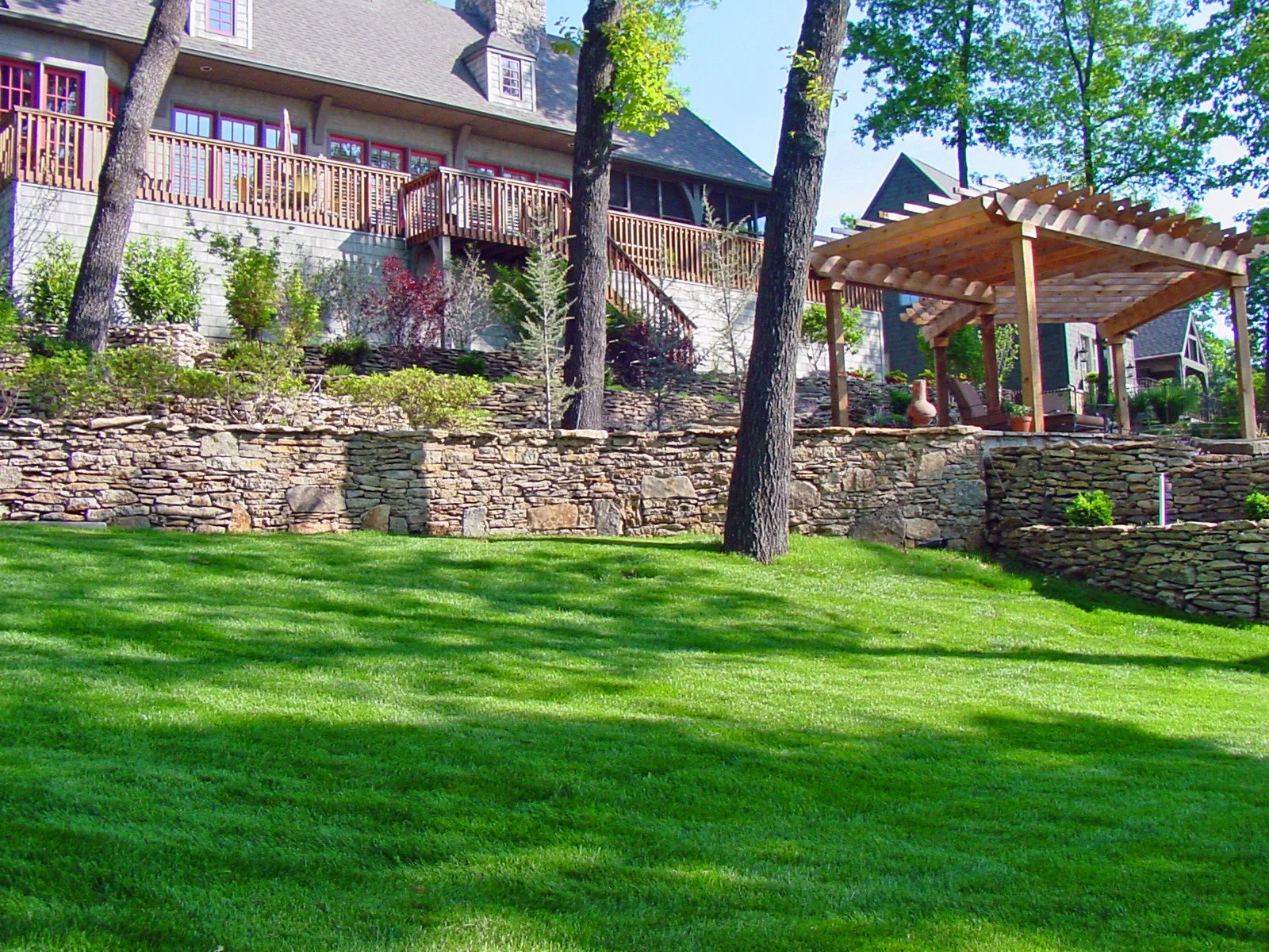 A landscaped backyard with a lush green lawn, stone retaining walls, tall trees, and a multi-level house with a wooden deck and pergola.