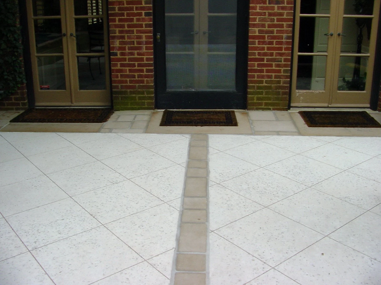 Front view of a brick building entrance with three sets of glass-paneled doors, black and beige, and three rectangular doormats. The center door is black, and the side doors are beige.