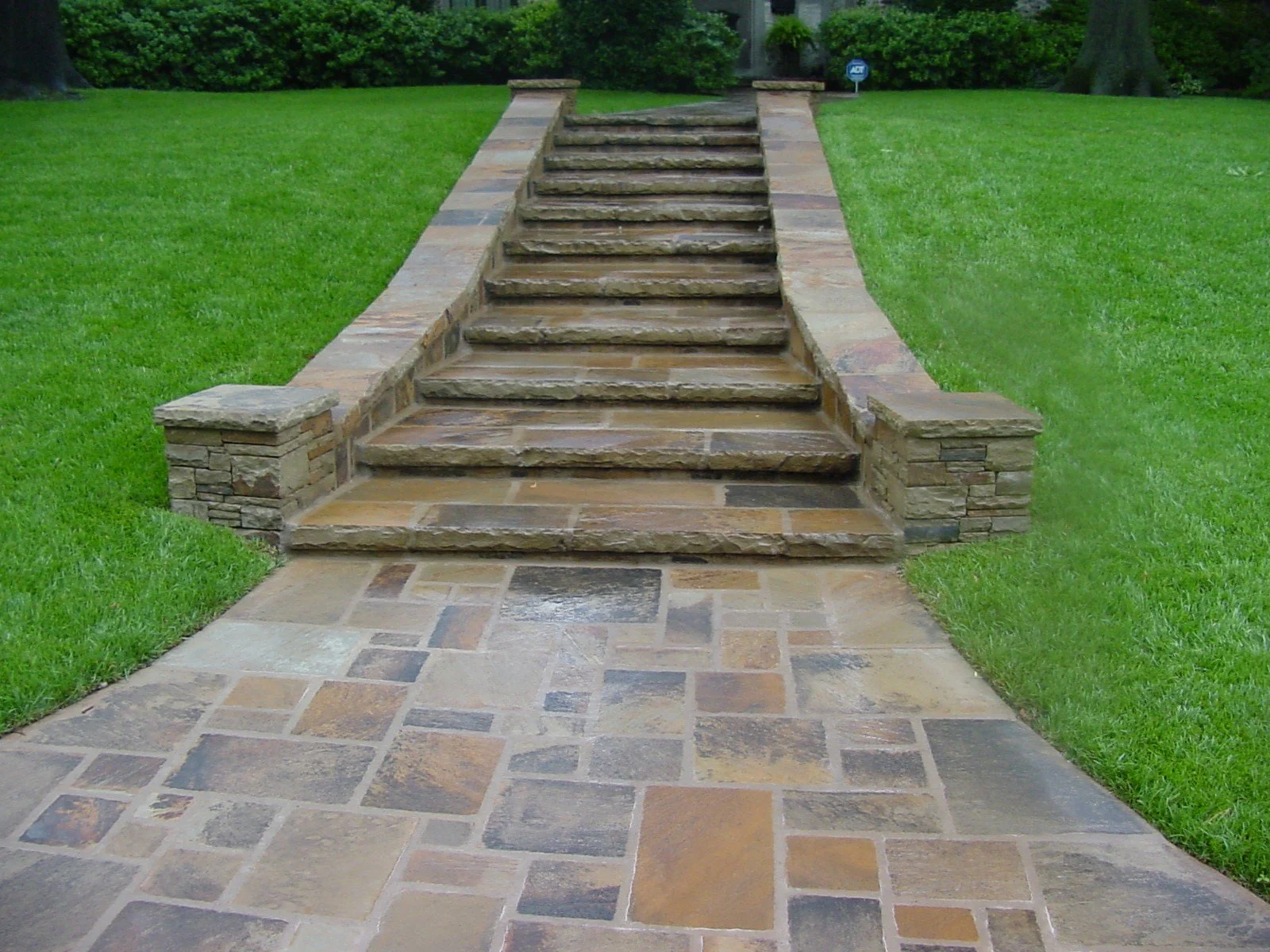 Stone and brick staircase leading from a paved walkway to a grassy lawn, with bushes and trees in the background.