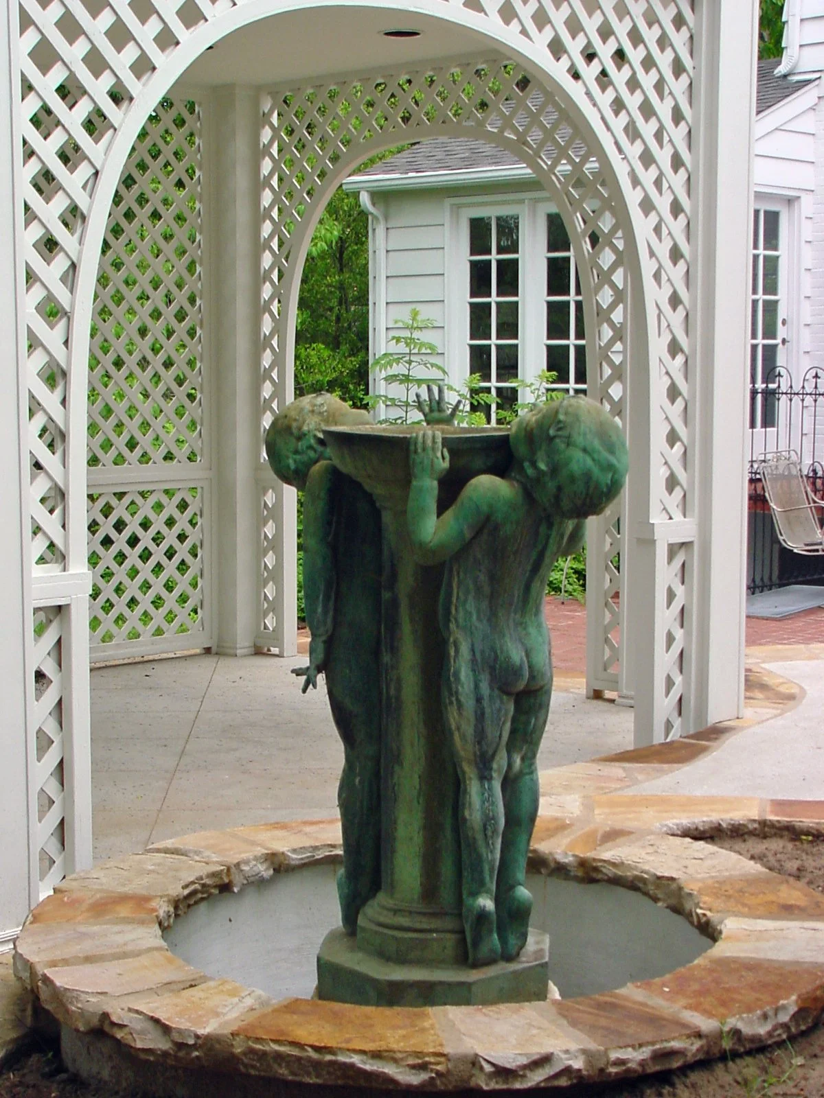 A greenish bronze sculpture of two children holding a large bowl, standing in the center of a stone fountain. The fountain is surrounded by a white lattice arbor and is located outdoors in a garden or patio area.