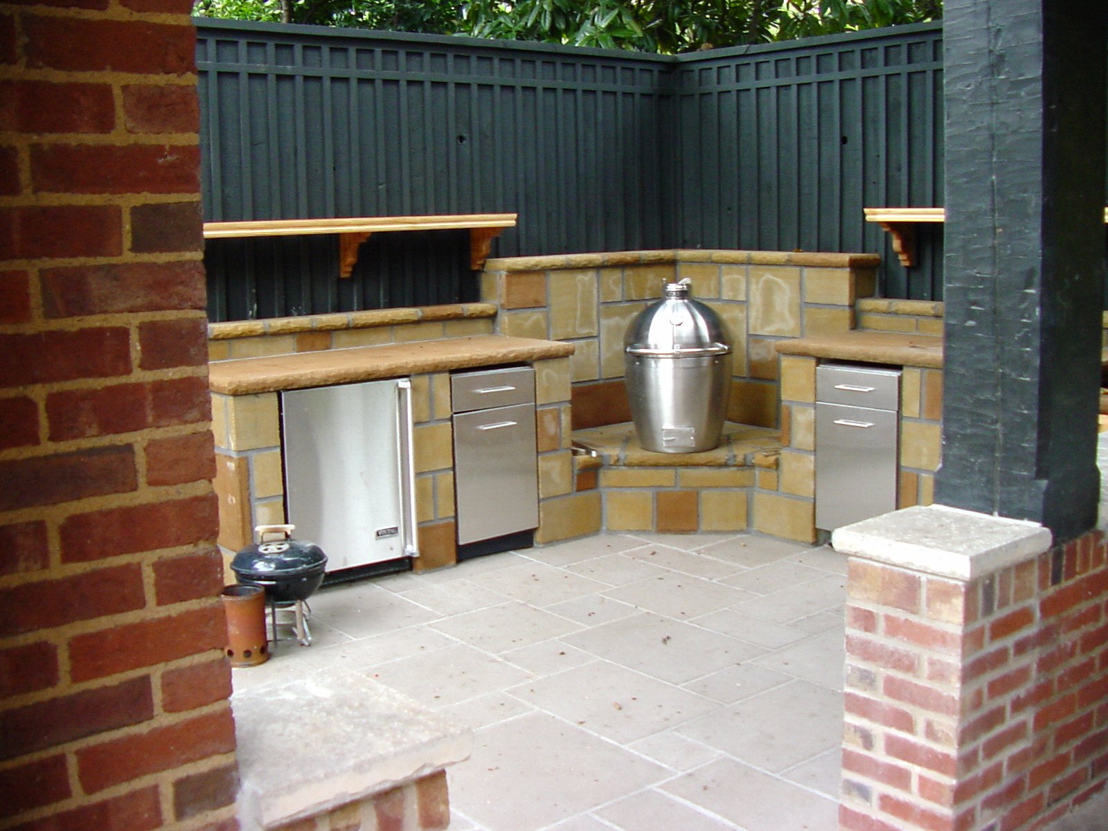 An outdoor kitchen area with a brick and tile construction, dark blue wooden fencing, and a small charcoal grill on the left. The countertop area includes small refrigerators and a large metal ice bucket in the center. There are brick pillars and a t