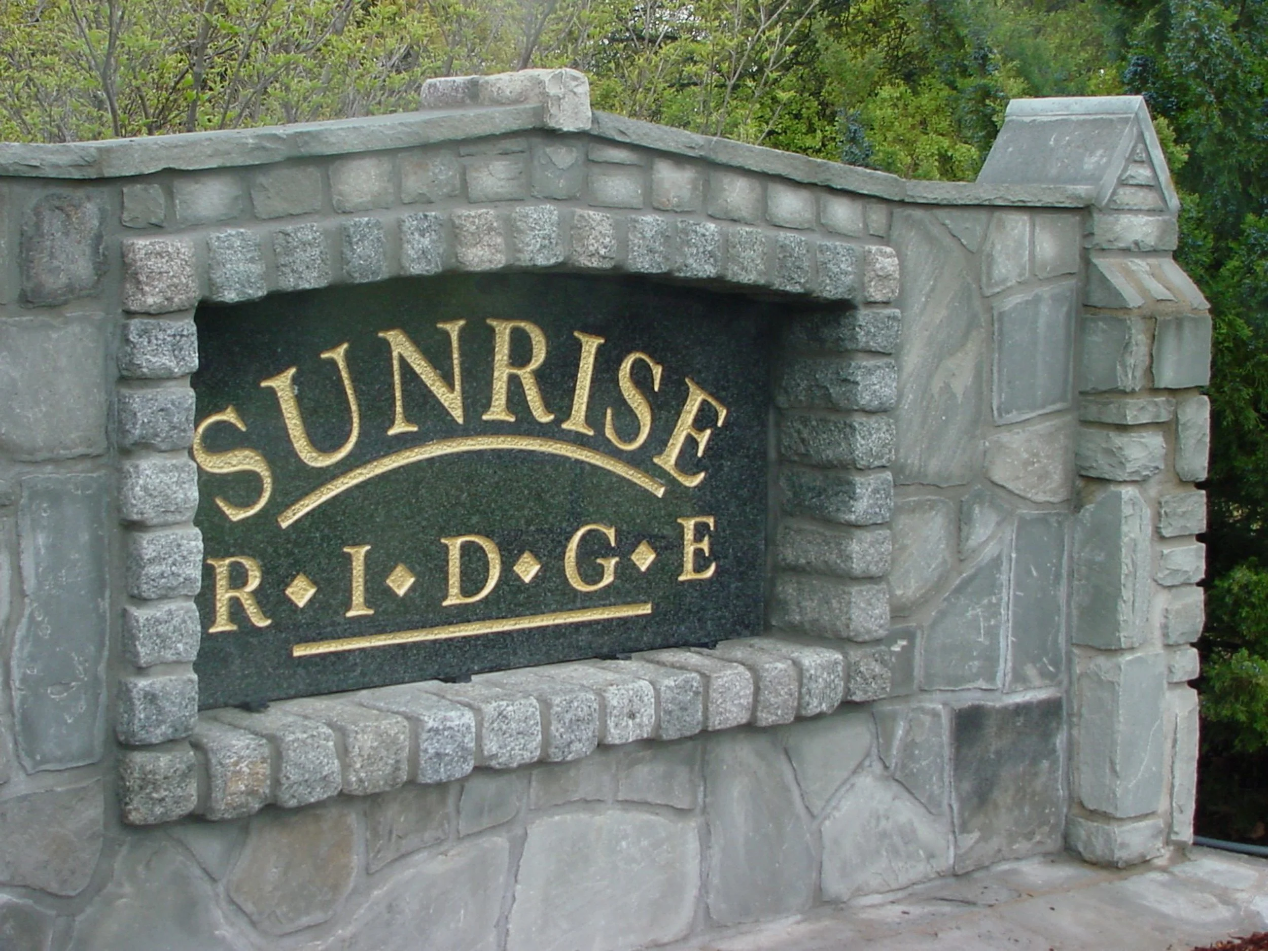 Stone sign with gold lettering reading 'Sunrise Ridge,' surrounded by greenery.