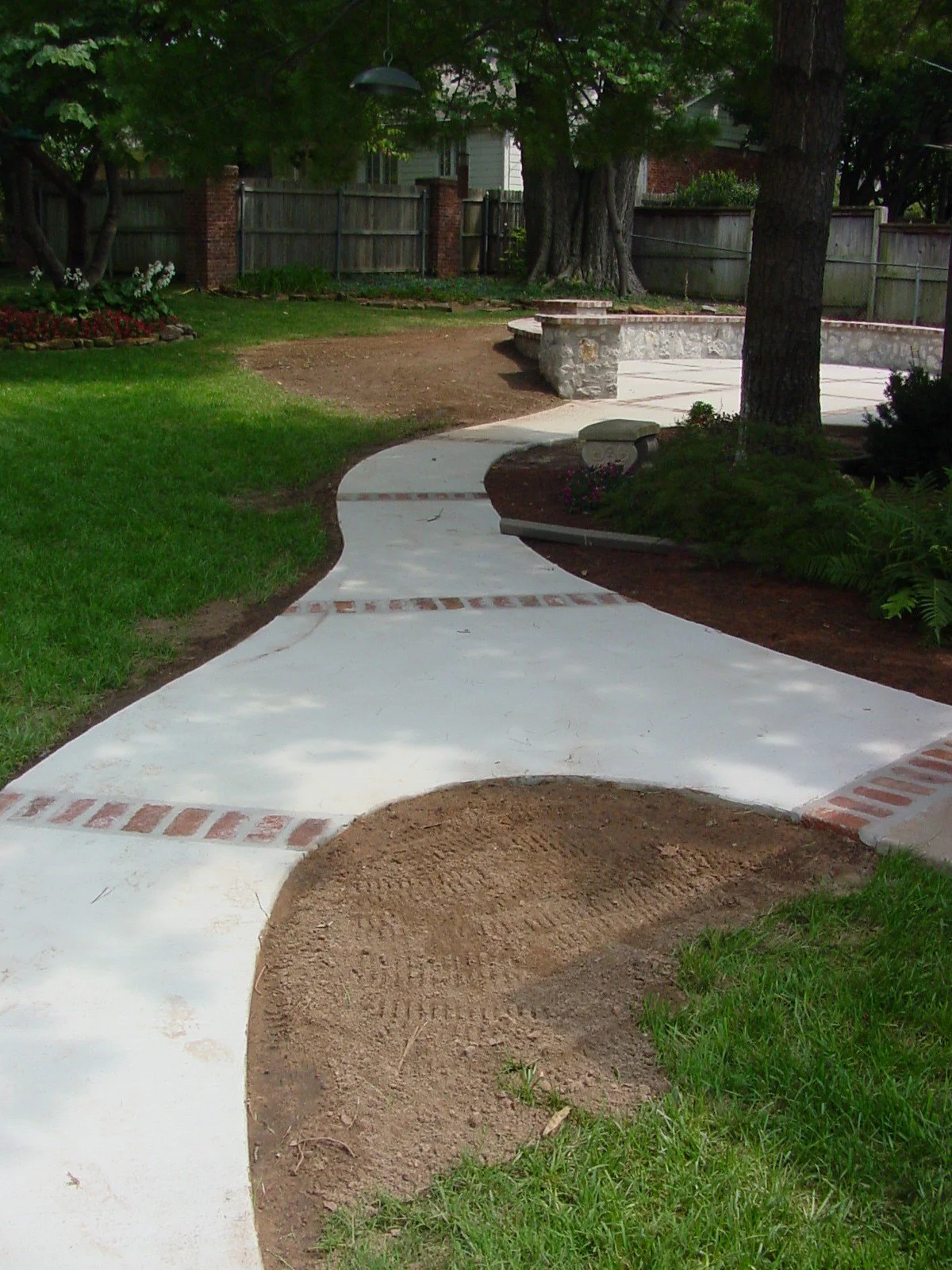 A winding concrete sidewalk in a backyard with a small garden bed on the left, green grass, and large trees in the background. The sidewalk has decorative brick accents and a section of dirt where it is being repaired.