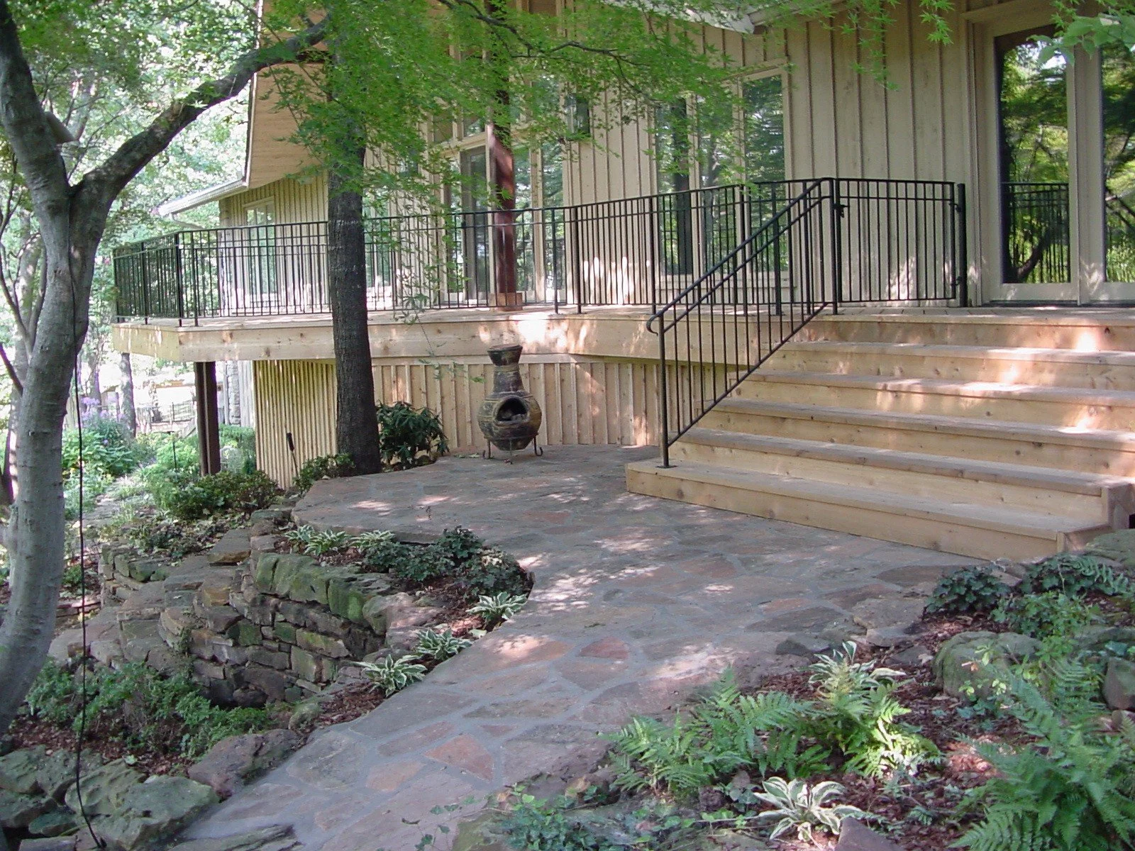 A backyard deck with a metal railing, wooden stairs, and a stone patio bordered by a stone wall with plants and shrubs. There are trees providing shade.