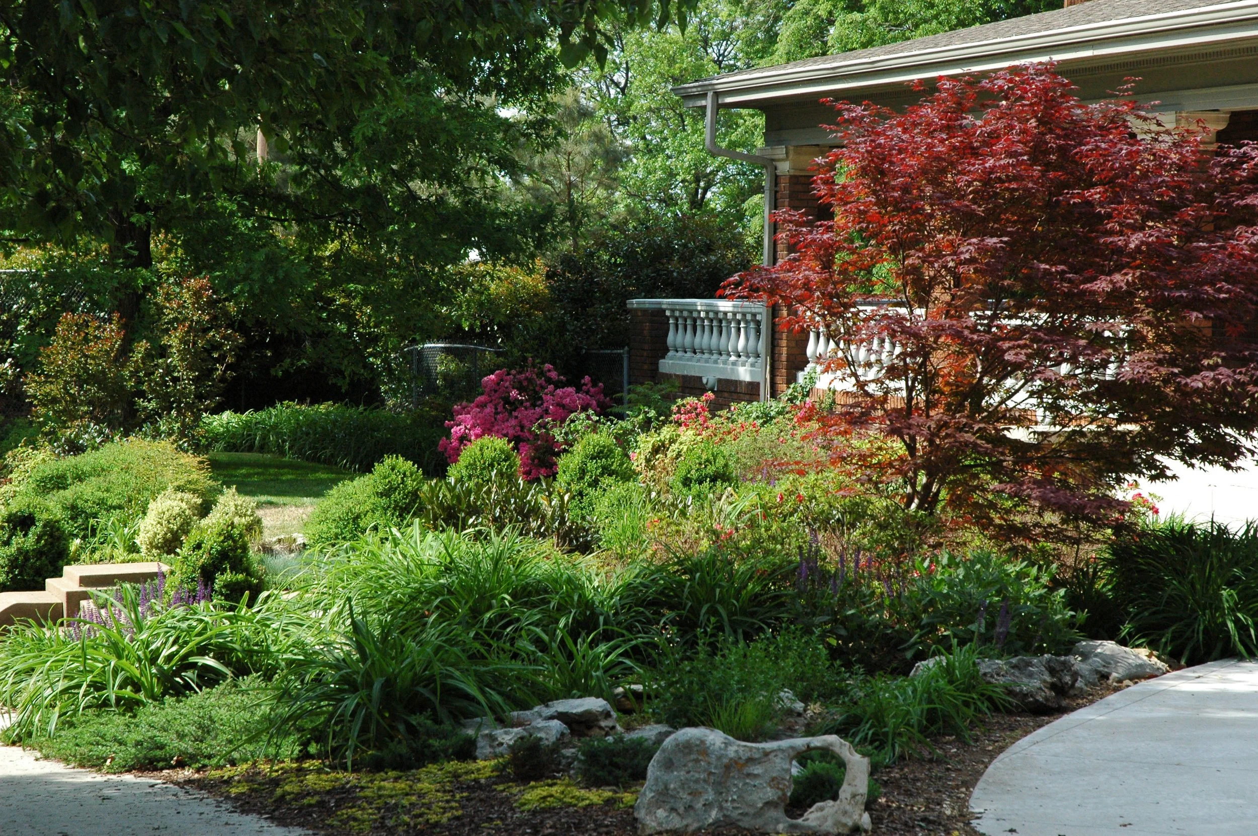 A lush, green garden with various plants and flowers in front of a house. The house has a porch with a white railing, red brick walls, and a gray roof. A red-leafed tree is prominent on the right side, and a curved concrete pathway runs through the g