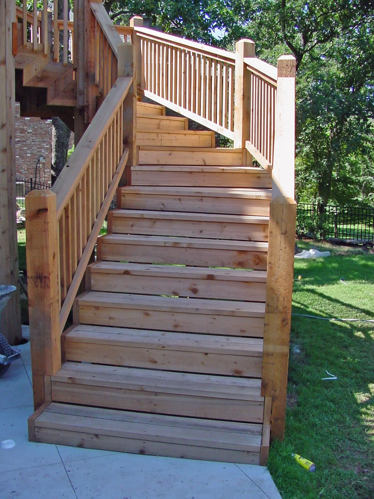 A newly built wooden staircase with railings leading from a patio to a yard, surrounded by green trees and grass.