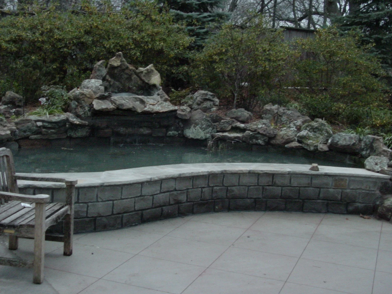 A small pond with a stone border and a waterfall feature surrounded by rocks and trees in a garden setting.