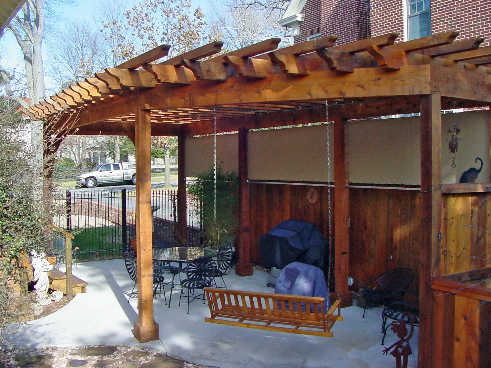 Backyard patio with a wooden pergola, outdoor furniture including a round table with chairs, a covered grill, and a bench swing. In the background, there is a brick building, a street with parked cars, and trees with bare branches.