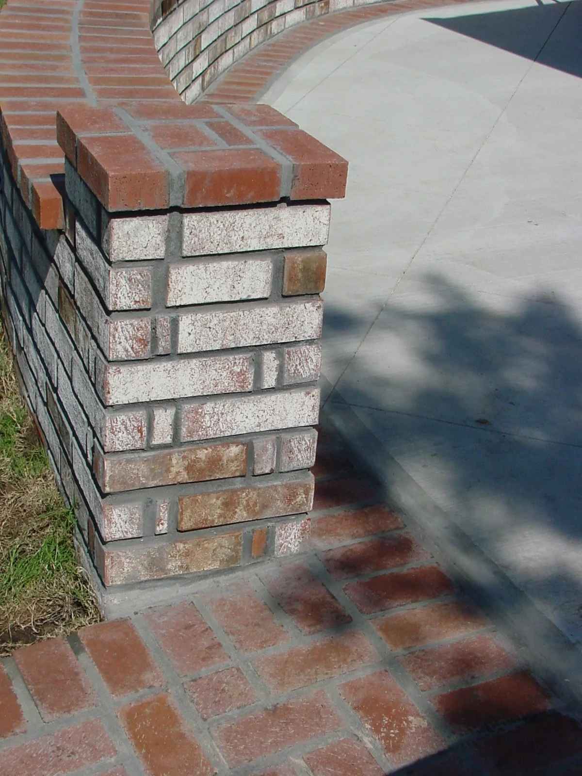 Close-up of a brick pillar with an arched brick structure beside a concrete sidewalk and a brick-paved area, with grass visible on the left.