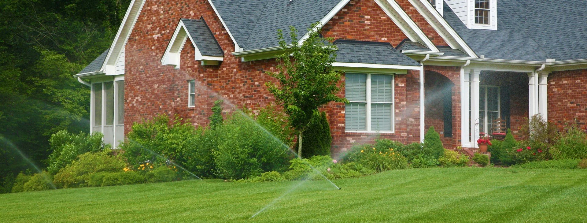 The front yard of a brick house with a lush green lawn being watered by a sprinkler system.