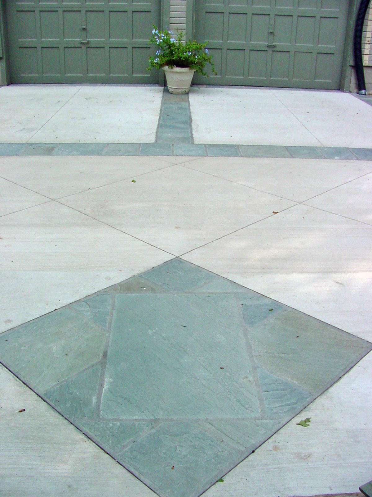 Concrete driveway with decorative green ceramic tile pattern, a large potted plant with foliage and flowers, and a closed green garage door in the background.