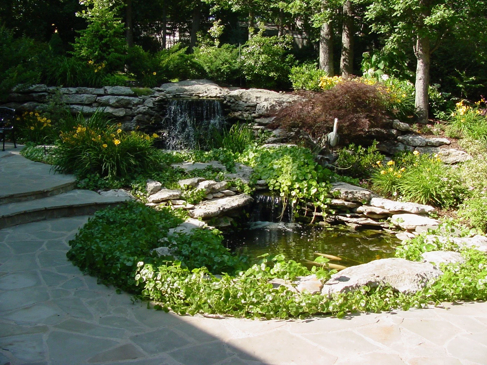 A landscaped garden with a small water fountain, lush green plants, and a stone pathway.