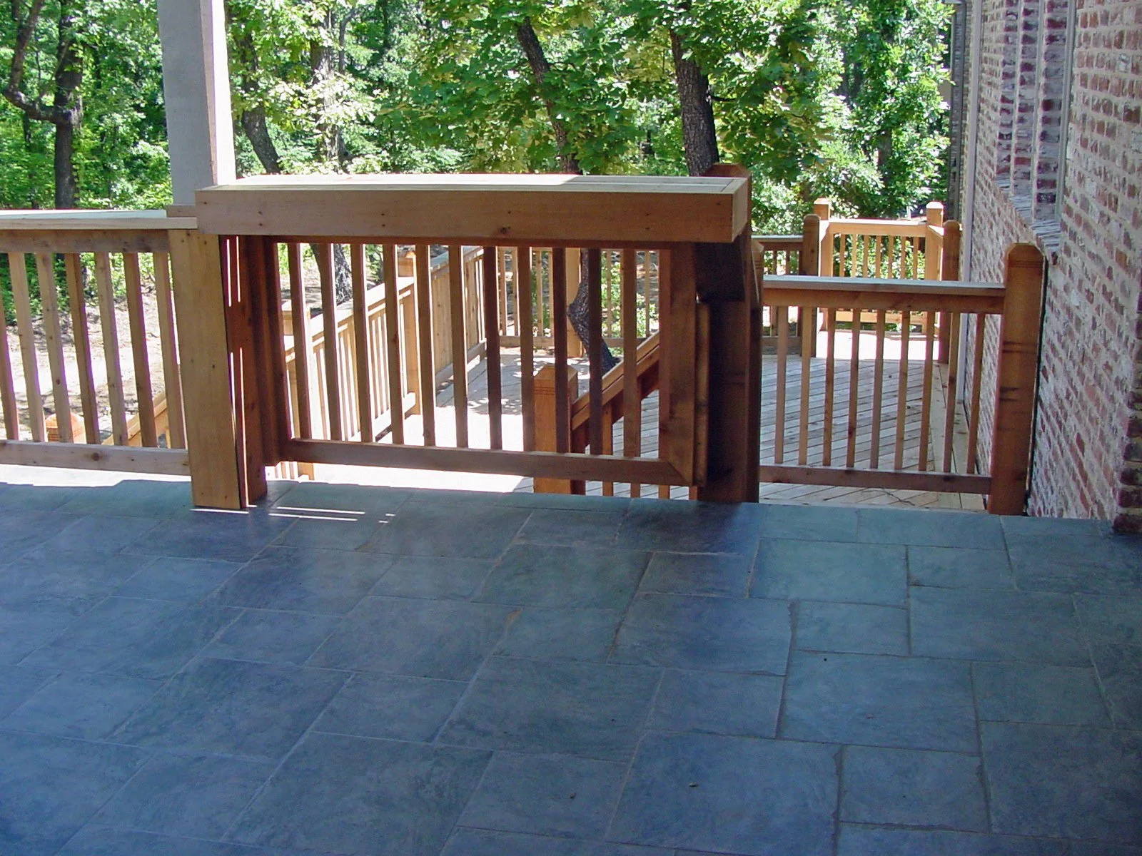 View from a porch showing blue tiled floor, wooden railing and stairs leading down to a yard with trees.
