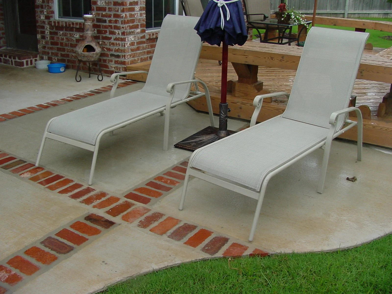 Two white outdoor lounge chairs on a concrete patio with red brick accents, next to a brick house, with a blue umbrella and a wooden patio deck with potted plants in the background.