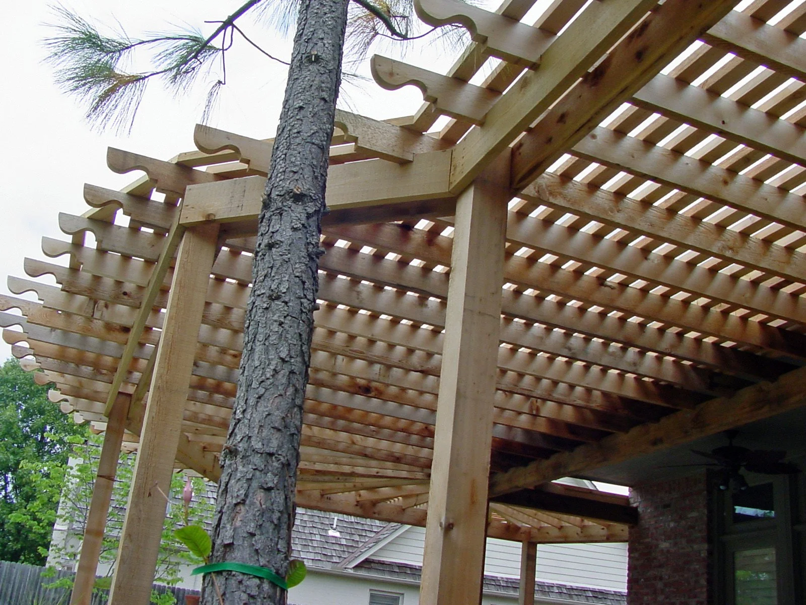 Wooden deck under construction with support beams, a tree trunk in the middle, and neighboring houses in the background.