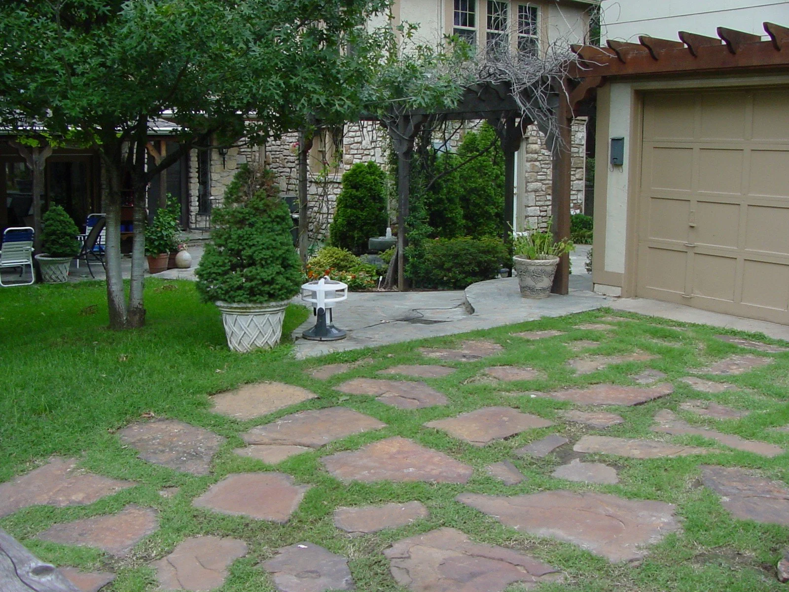 A landscaped backyard with a stone pathway, green grass, trees, potted plants, and a brick house with a beige garage door.