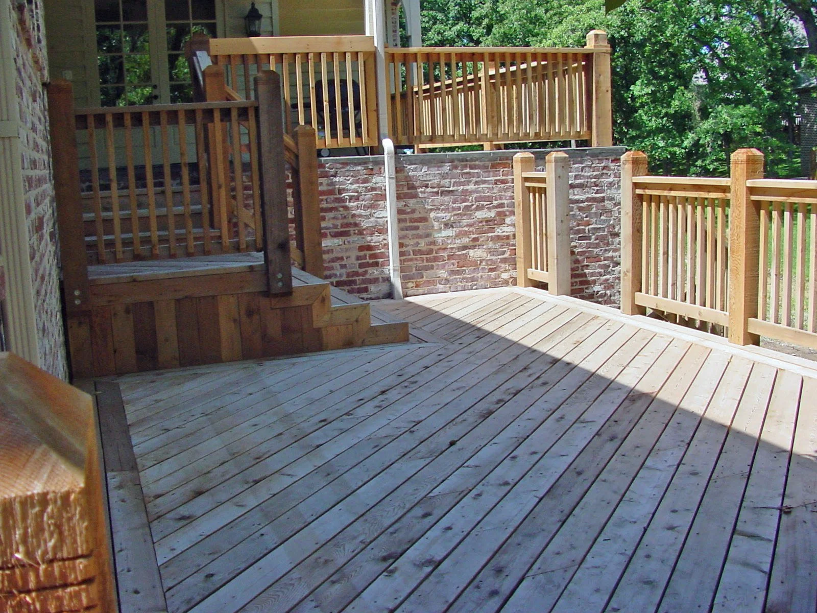A multi-level wooden deck with stairs, surrounded by wooden railings and a brick wall, with green trees in the background.