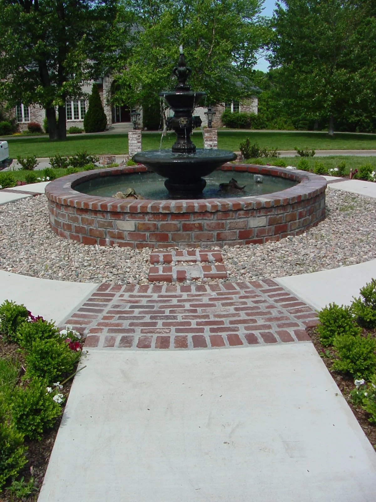 Brick and stone fountain with water flowing into a pool, surrounded by a brick circular wall and gravel, with a brick and concrete pathway leading to it, greenery, trees, and a large house in the background.