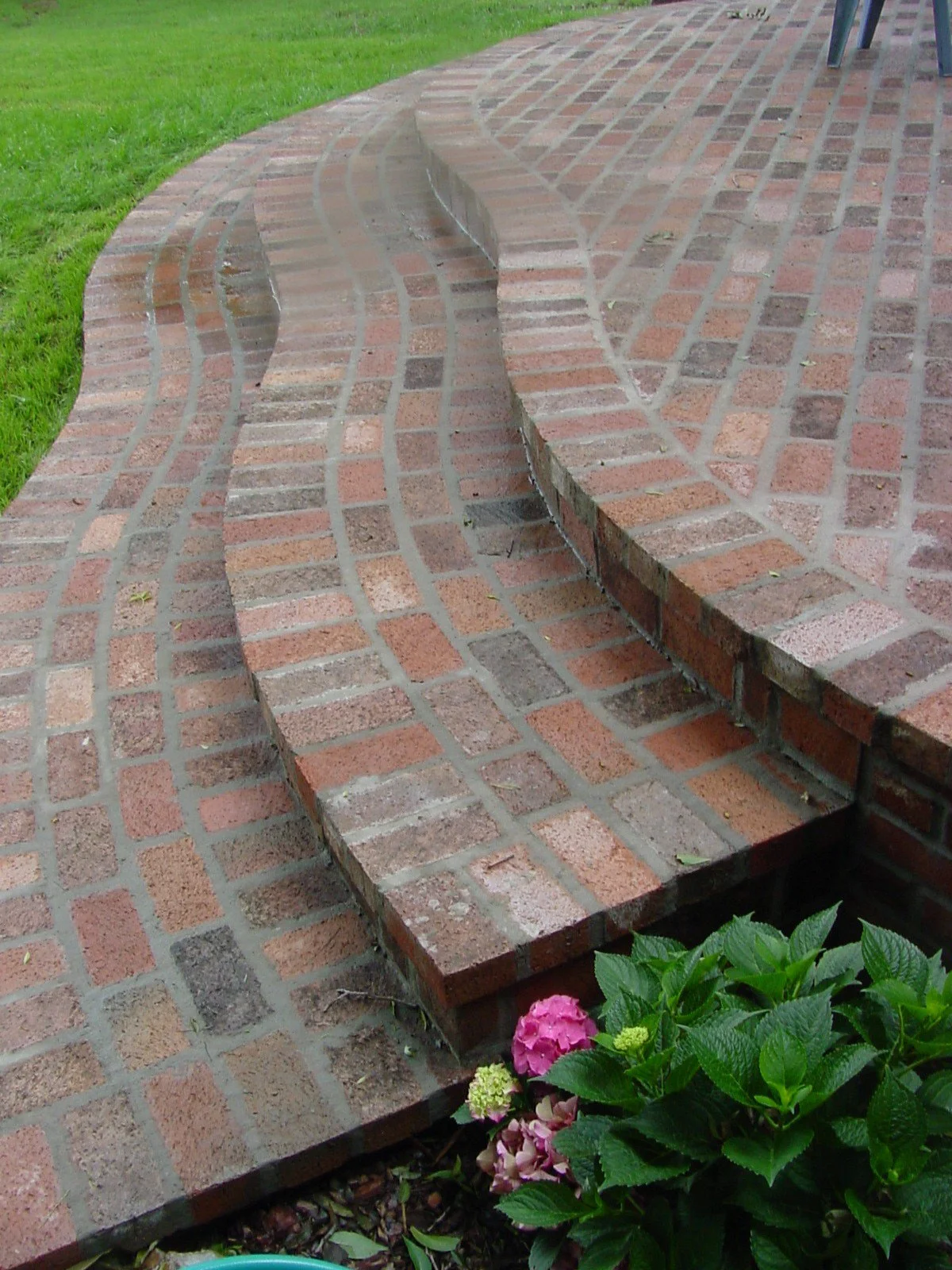 Decorative brick pathway with curved steps, green grass on the side, and flowering plant with pink and purple blossoms at the front.