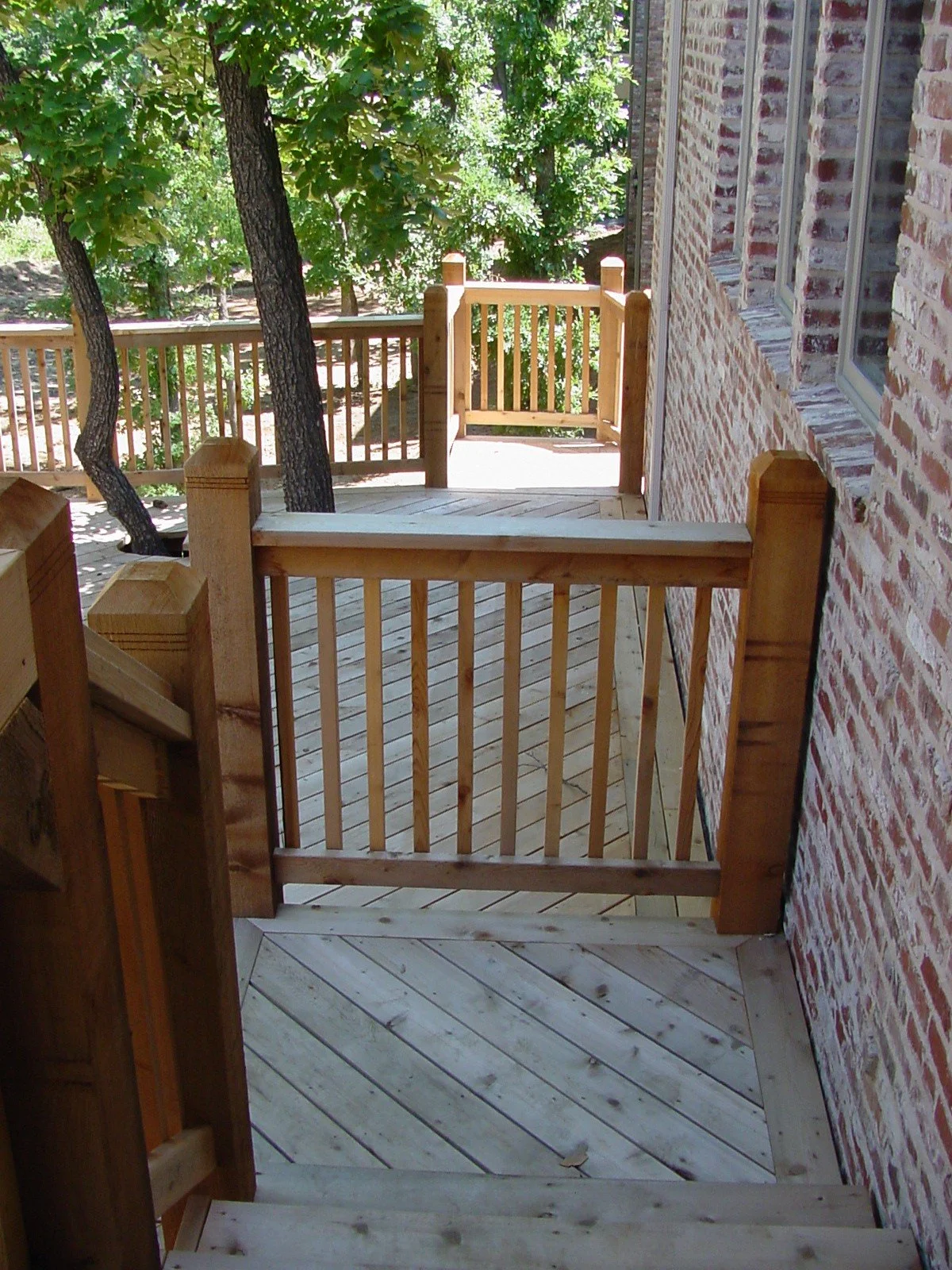 Back porch with wooden railing and steps next to brick wall, surrounded by green trees.